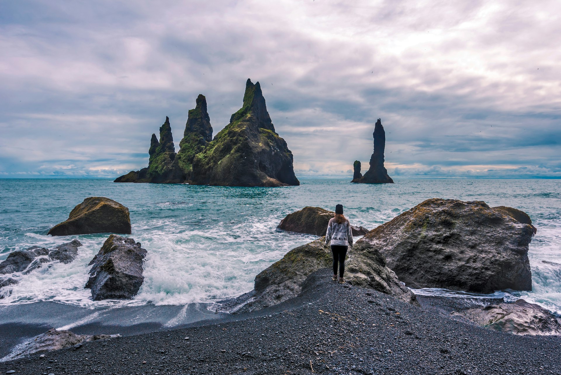 A girl approaches the rocky shore and basalt sea stacks at Reynisfjara beach.