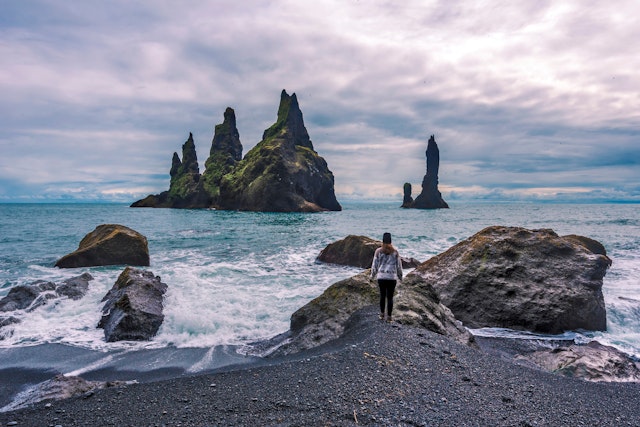 A woman stands on the rocks of a wild beach with several jagged stacks out at sea