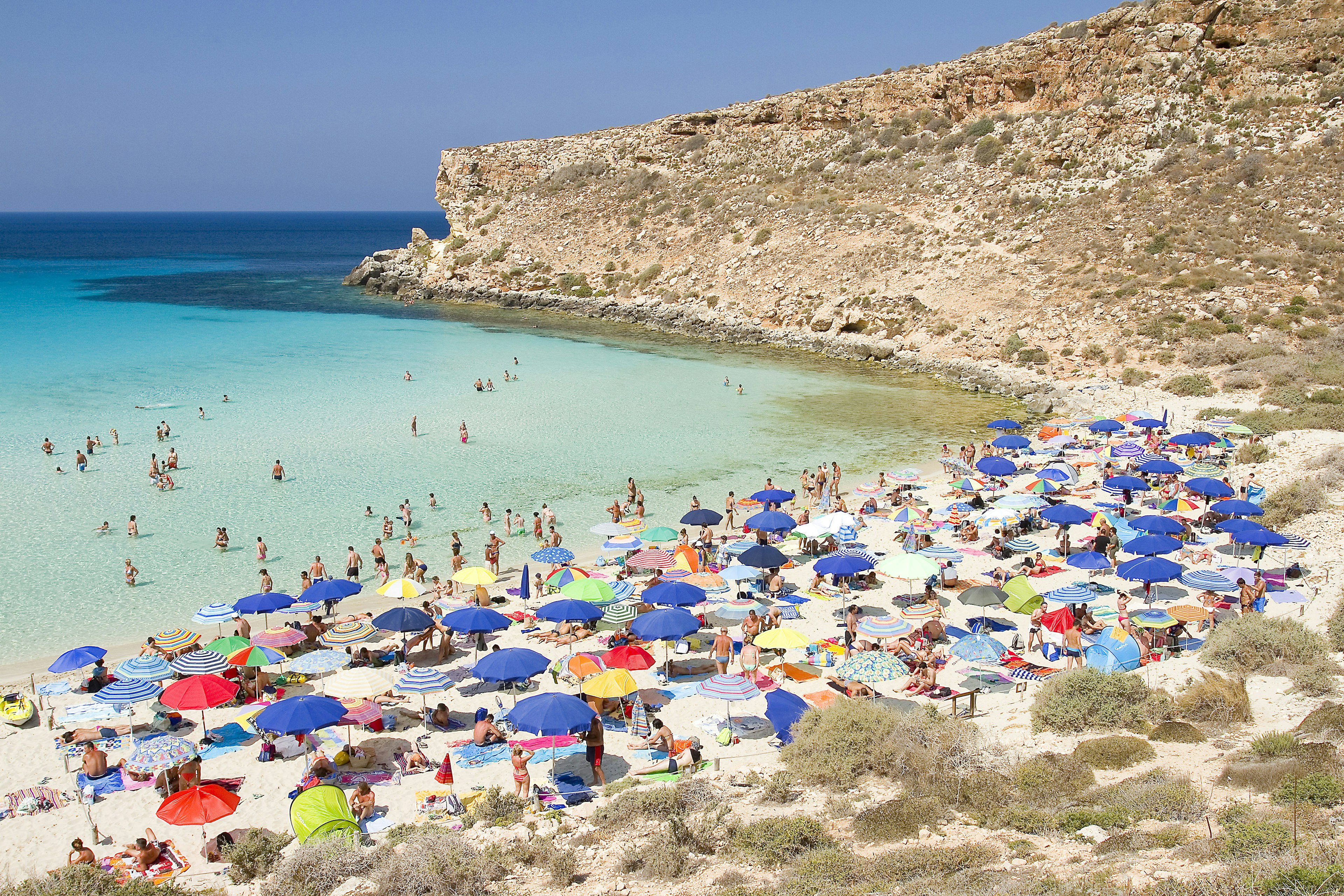 Tourists crowd a small beach, with colorful sun shades