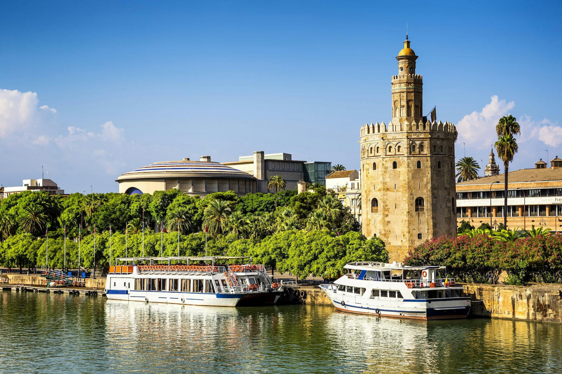 Seville's Torre del Oro, a circular stone tower with a minaret next to the river; white boats are moored in front of it and there are trees and bushes on either side of the tower.
