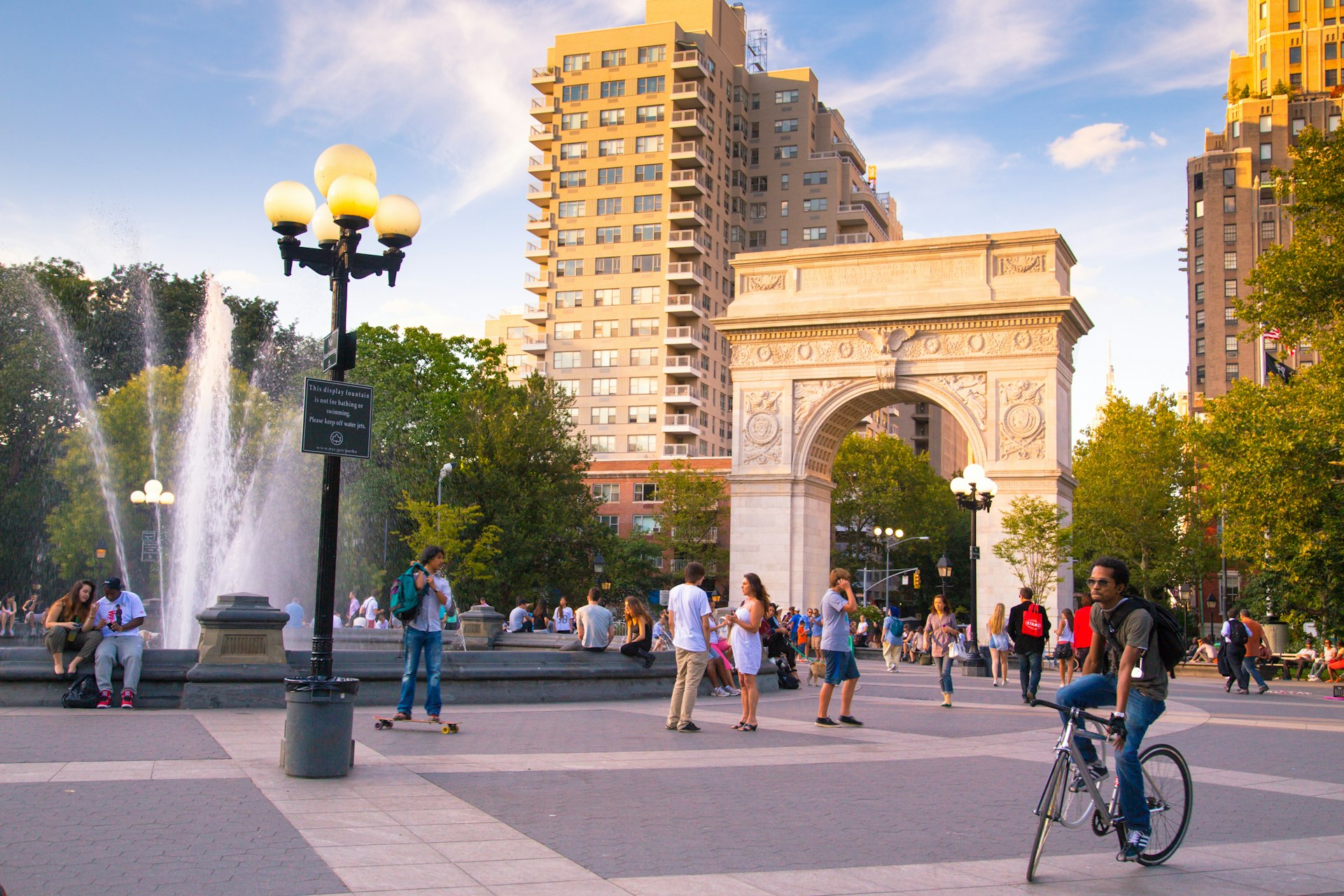 People walk and a person rides a bike during a summer evening at Washington Square Park in Manhattan