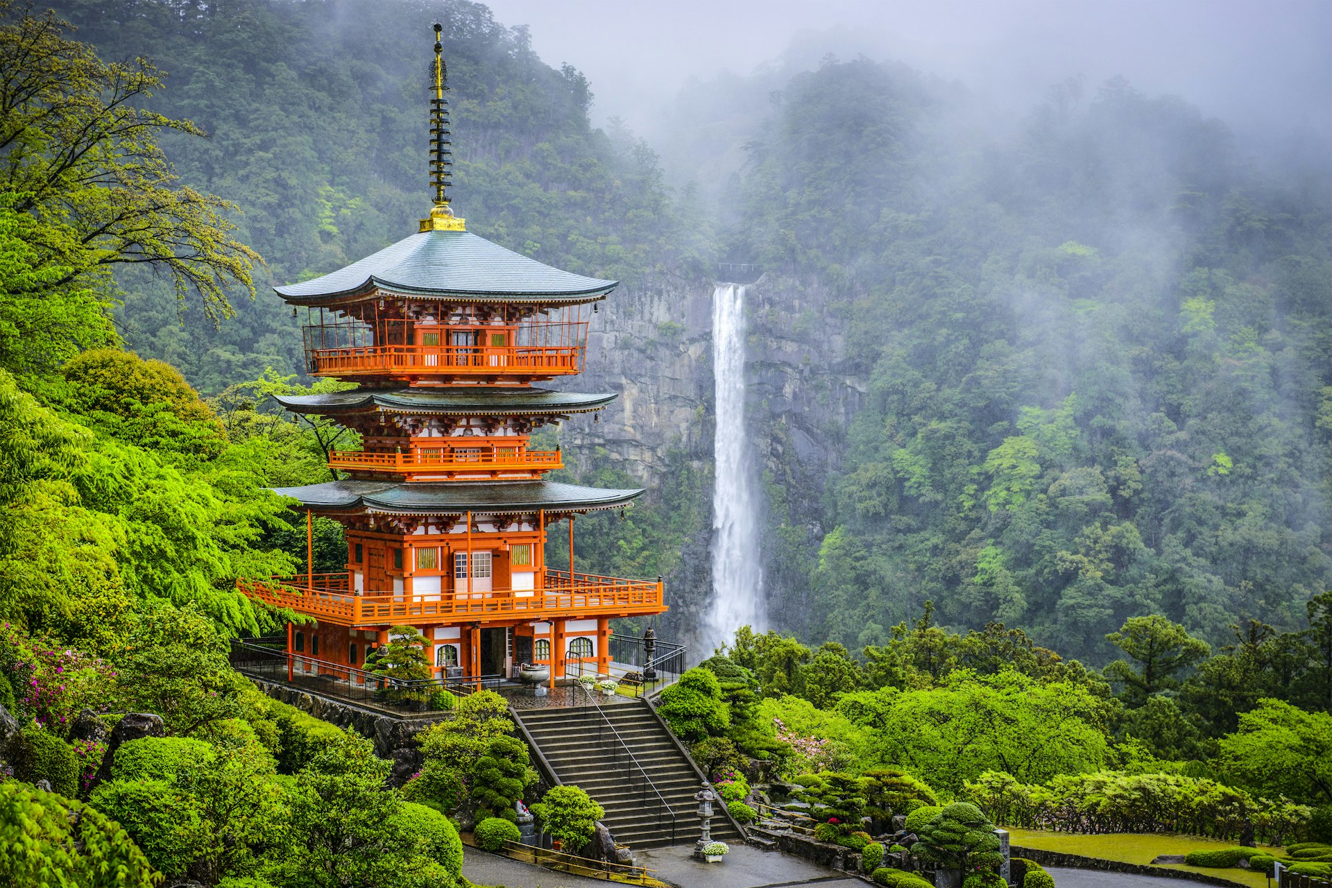 A red-roofed many tiered pagoda in the foreground, with a narrow, tall waterfall dropping amongst greenery in the background. Mist is descending over the hills