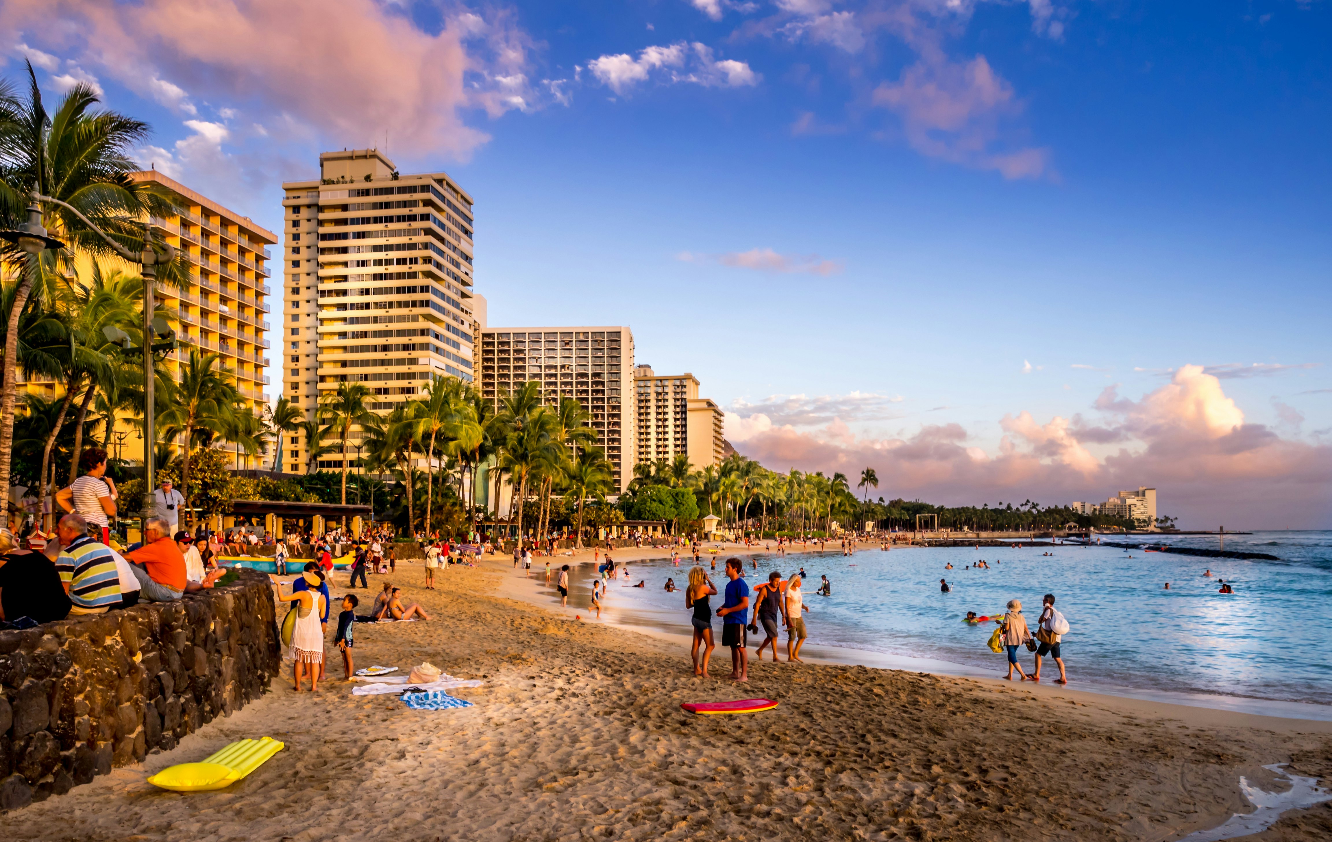 People on a sandy beach and in the water at sunset; tall buildings line the shore.