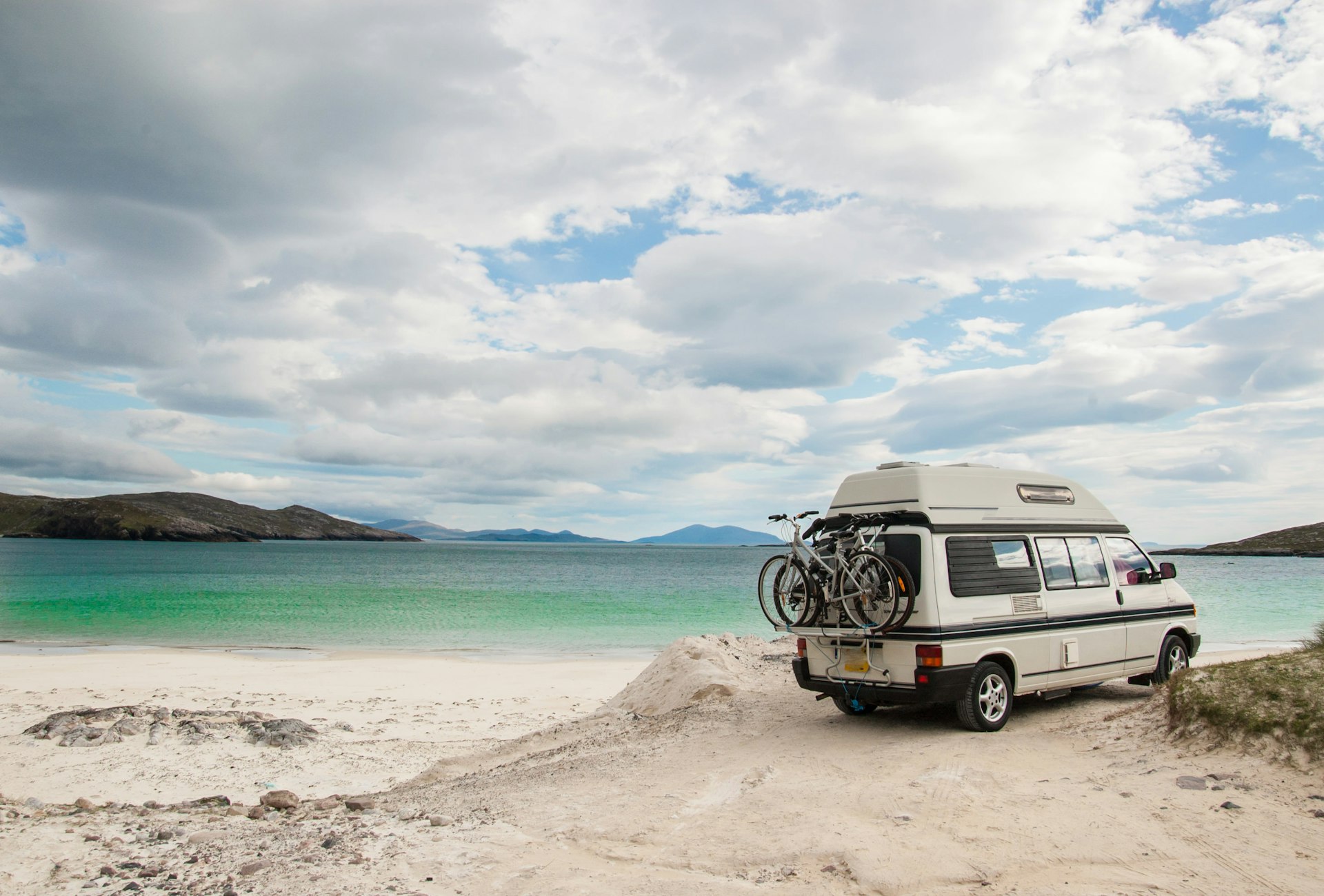 A blue VW campervan with two bikes on the back parked on a beach in Scotland