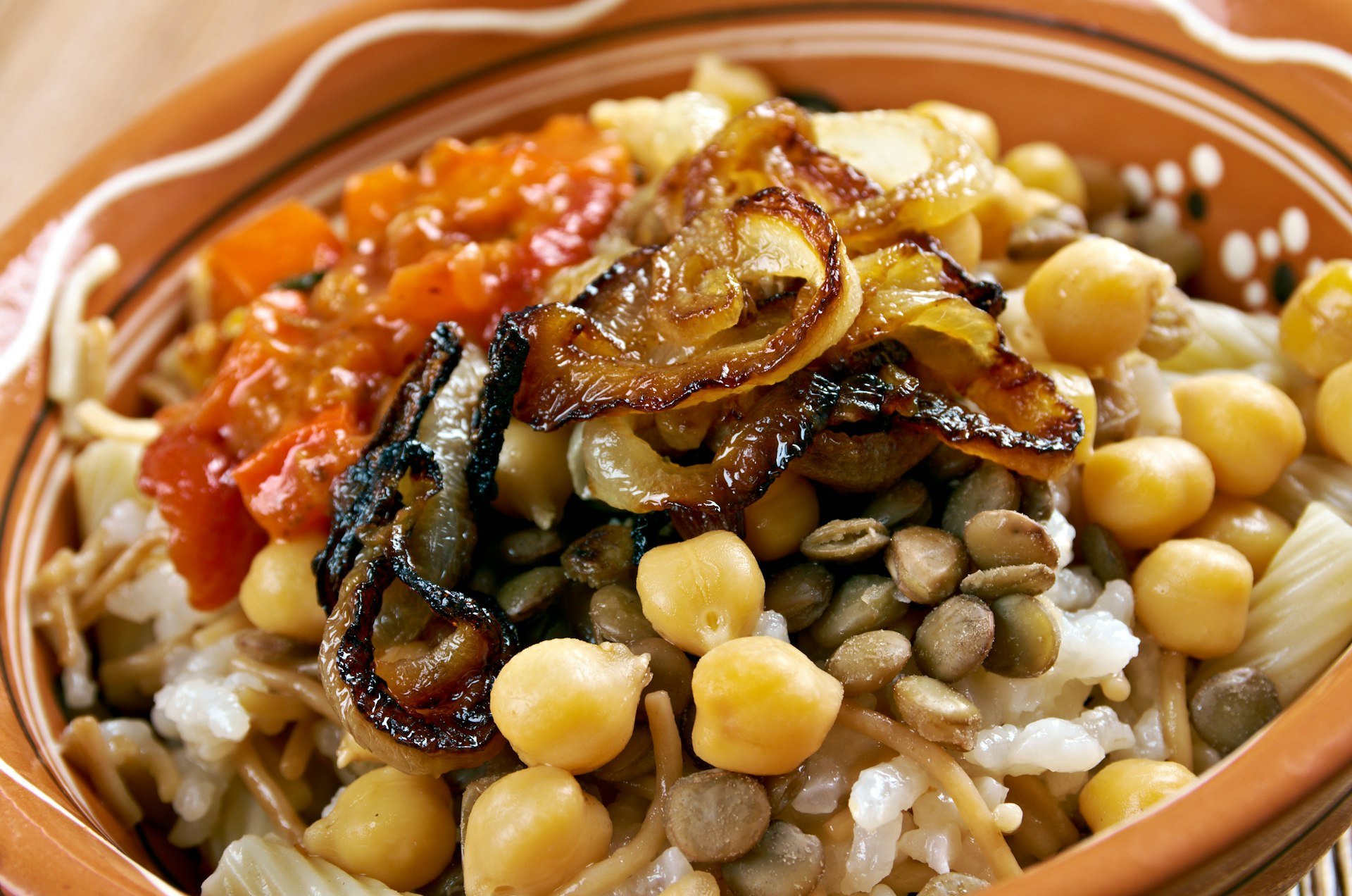 En mycket närbild av en skål med Kushari - en egyptisk maträtt med ris, makaroner och linser blandade, toppad med en tomatvinägersås;  några lägger till korta spagettibitar garnerade med kikärter och knaperstekt lök.