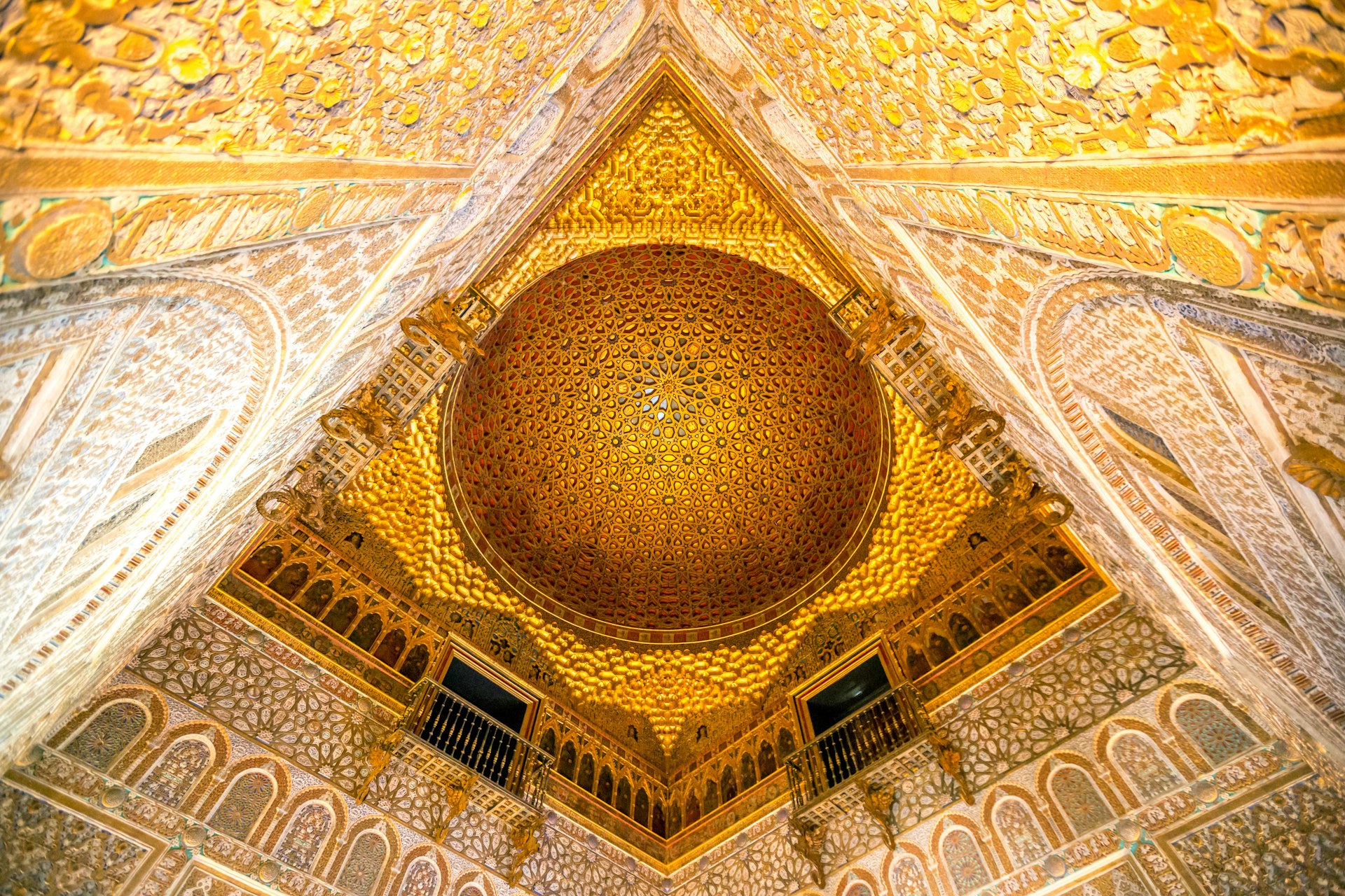 A view from below of an elaborate gold room in Seville's Real Alcázar; the walls and domed ceiling are covered in ornate tiles and mosaics.
