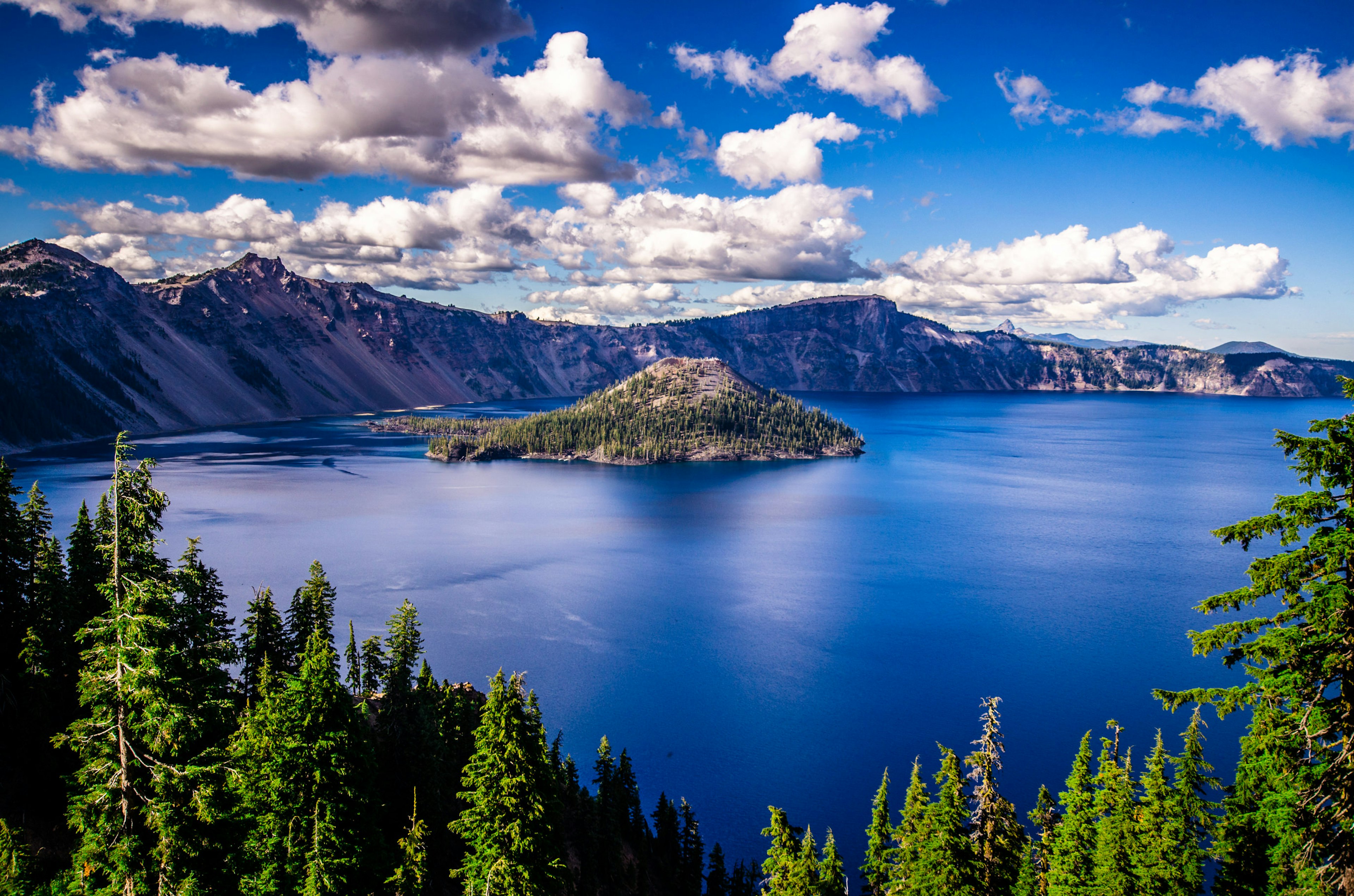 Aerial shot of dark blue waters of Crake Lake surrounded by thick green trees in Oregon. There are patches of clouds in the sky.