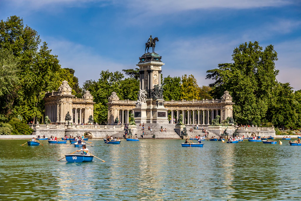 The Monument to King Alfonso XII, a grand colonnade that overlooks a central lake in Madrid's Parque del Buen Retiro, which has people in blue rowing boats on it.