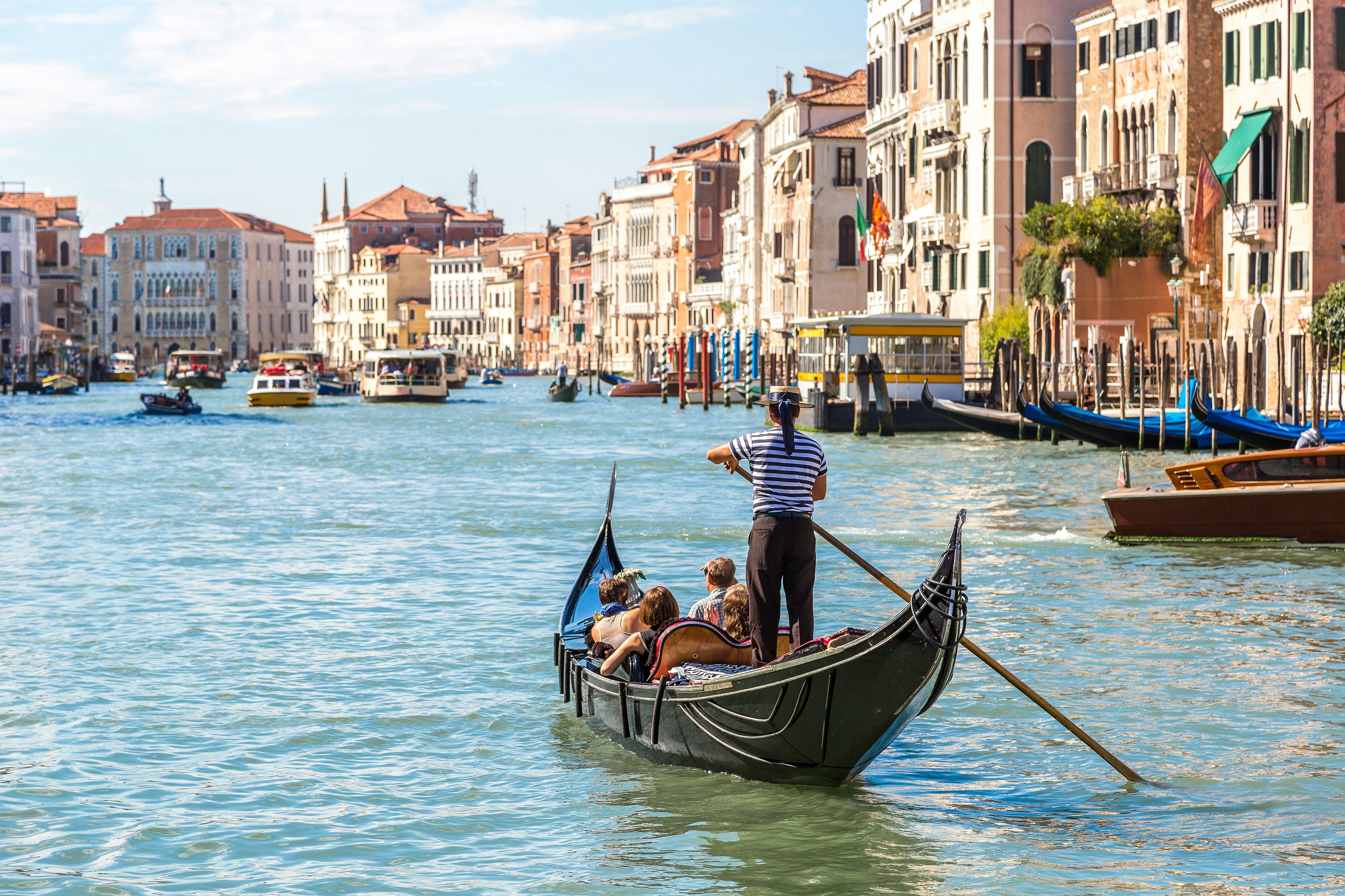 A gondolier steers a small black boat along a canal lined with historic buildings on a sunny day