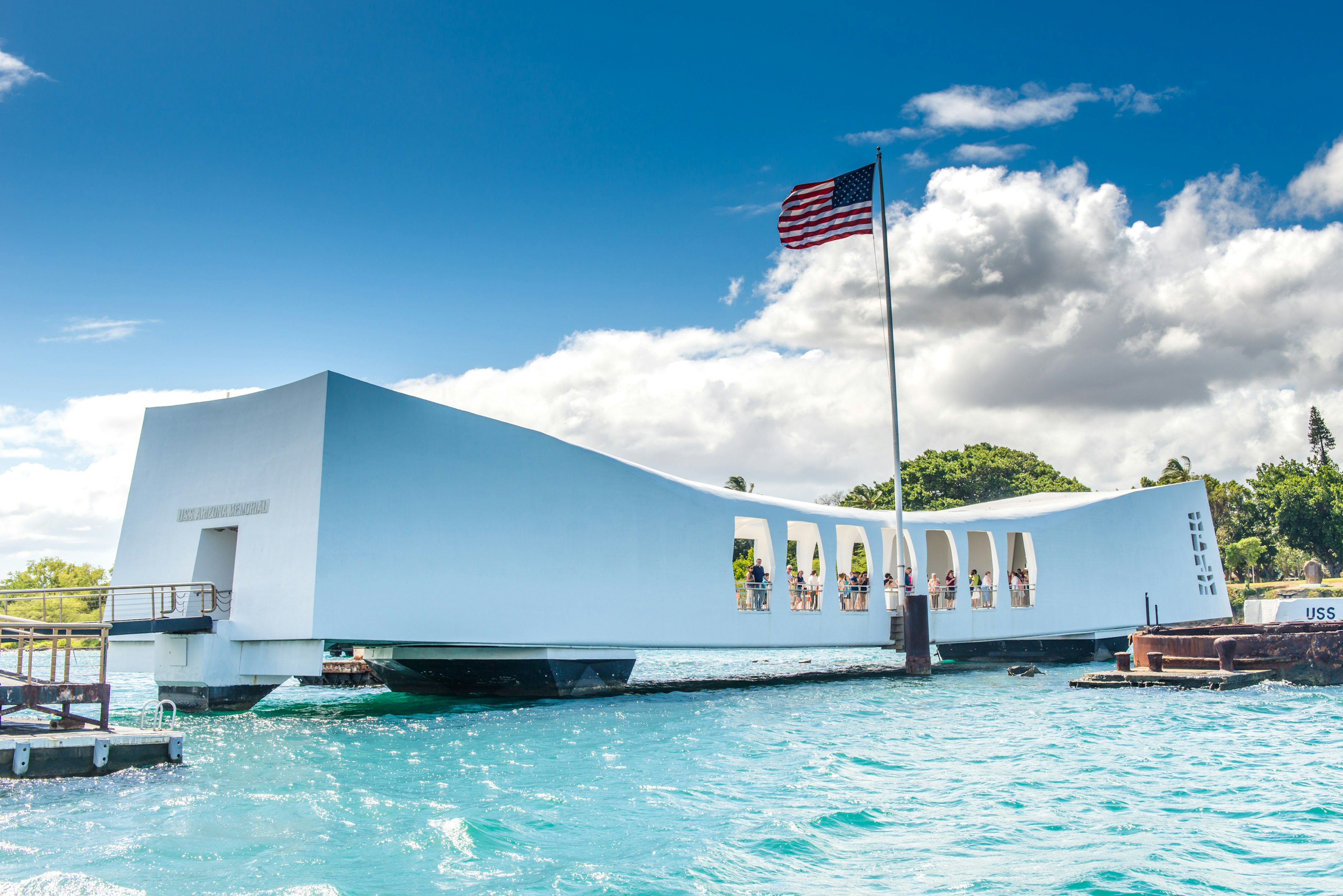 A white low-rise structure built over a waterway in a harbor. People are within looking at displays