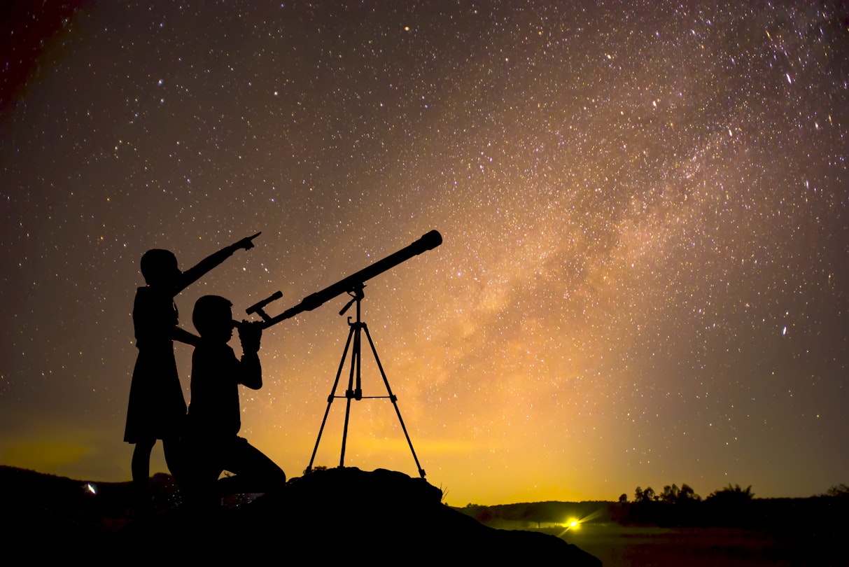Silhouette of children looking through a telescope at the Milky Way galaxy.