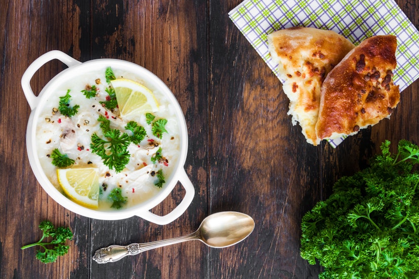 Avgolemono, chicken soup with egg-lemon sauce, rice and fresh parsley leaves in white bowl and freshly baked bread on wooden table. Top view.