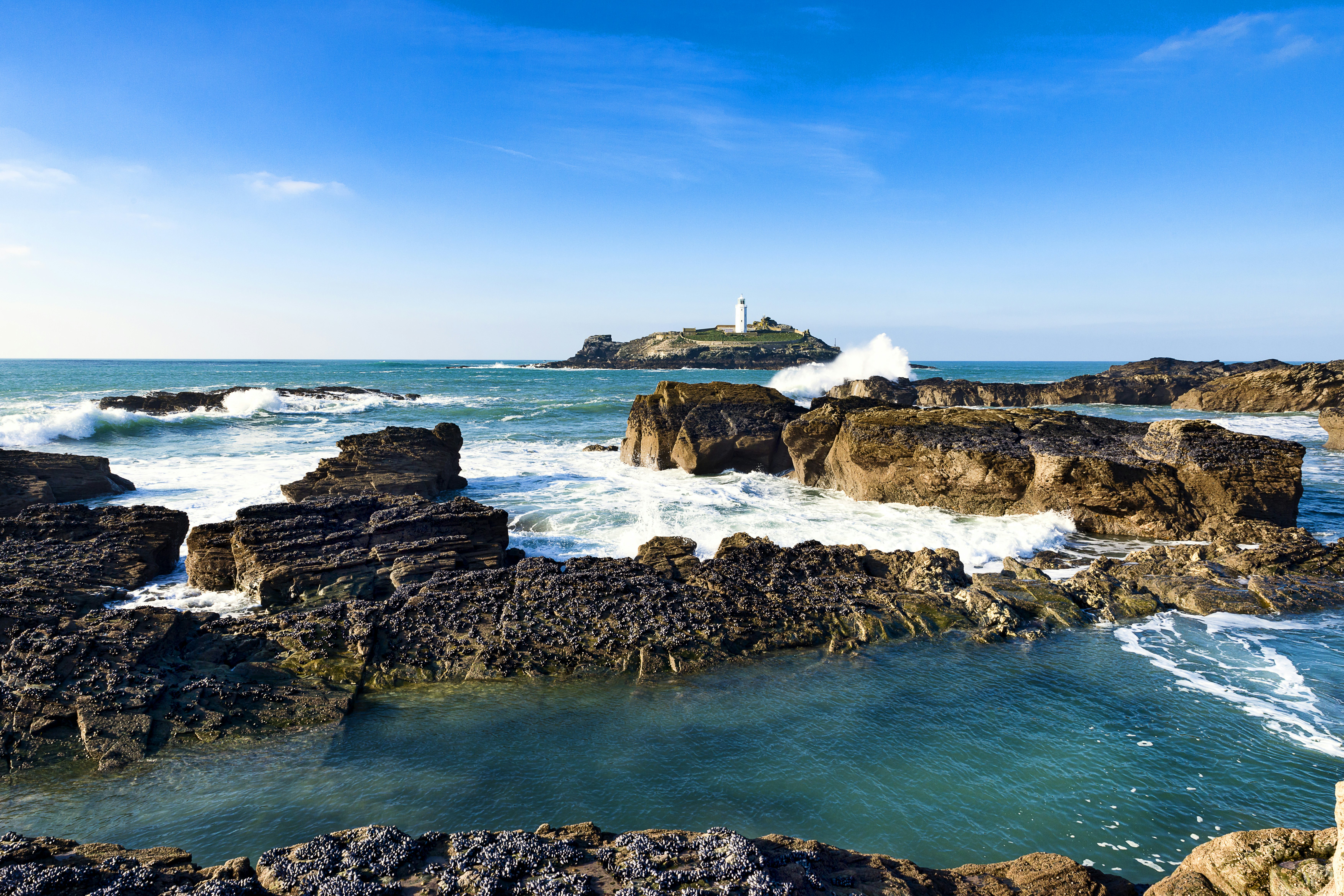 A lighthouse stands out on an island at sea. The shore is rocky with large waves