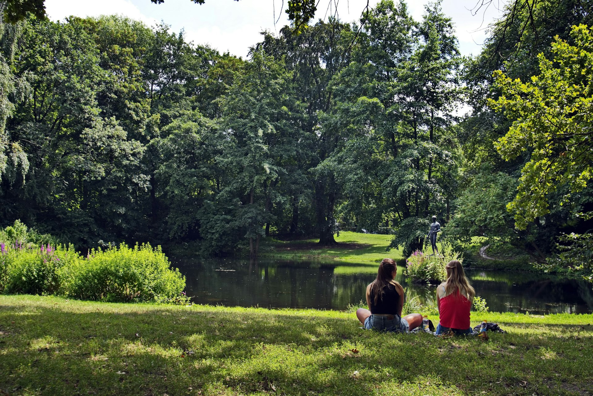 Two woman sit on the grass by a lake in Berlin park Tiergarten