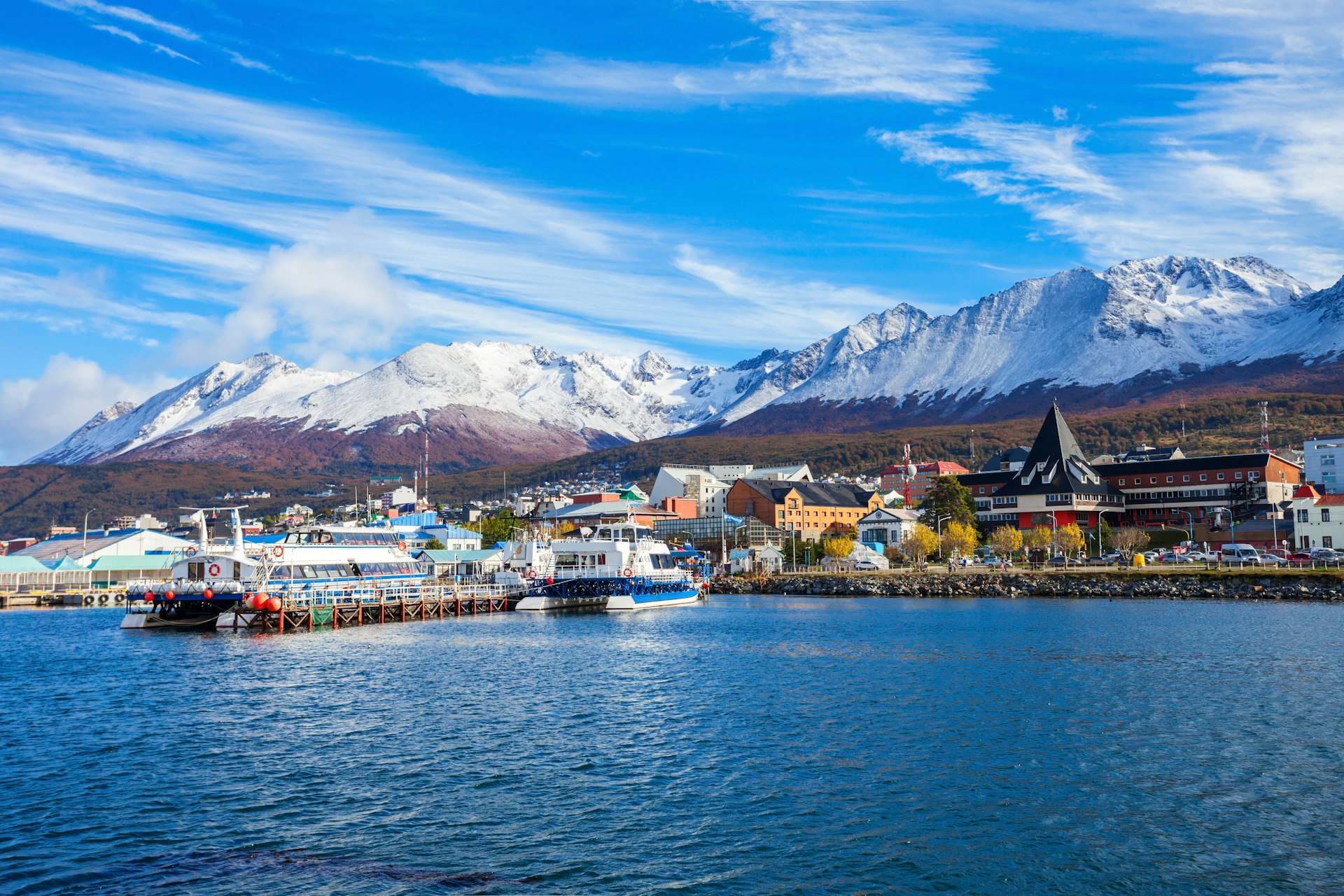 A small harbor lined with boats and catamarans; snow-capped mountains rise behind the town