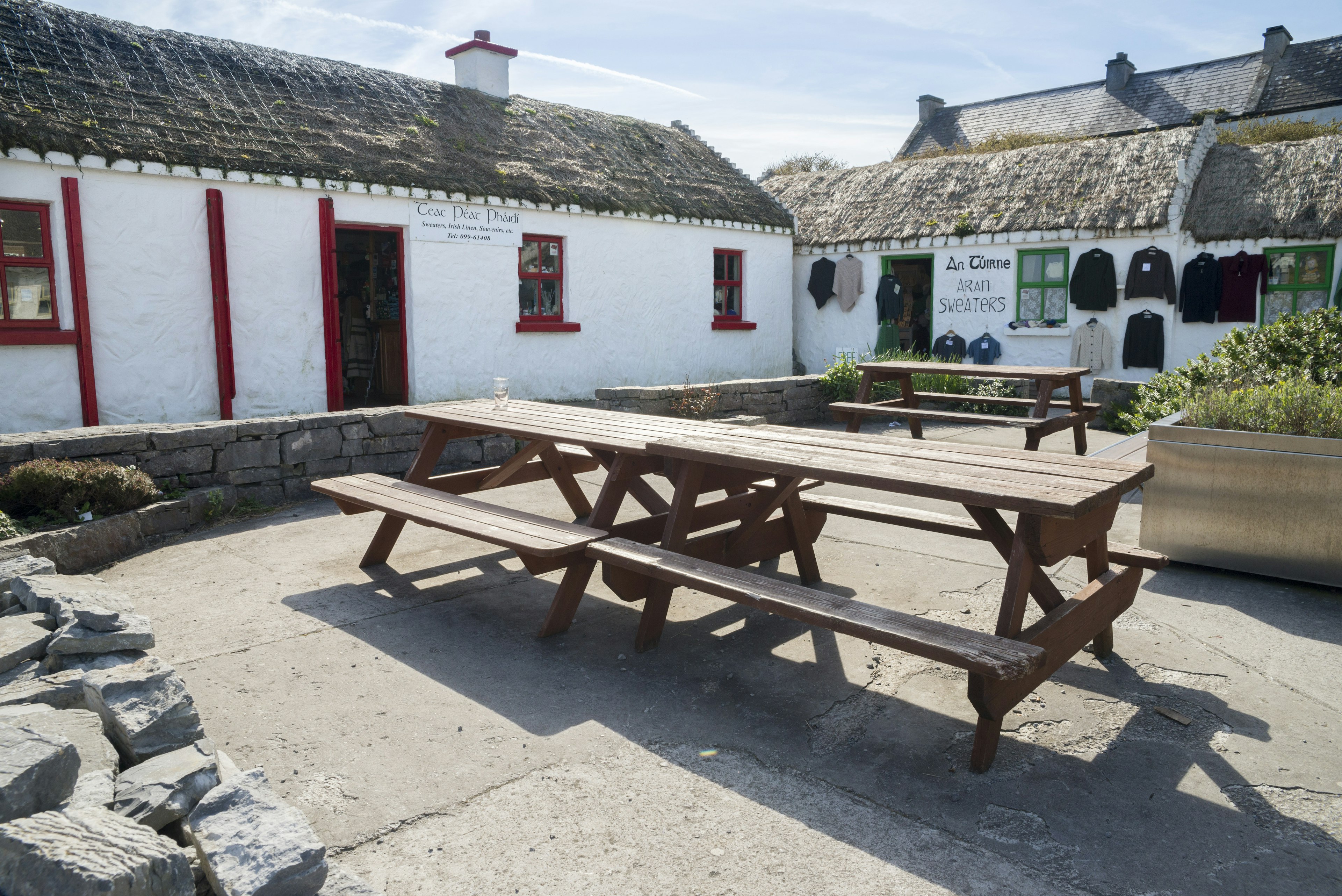 A low-rise white cottage with a thatched roof and picnic benches outside. Aran sweaters are for sale, hanging on one wall