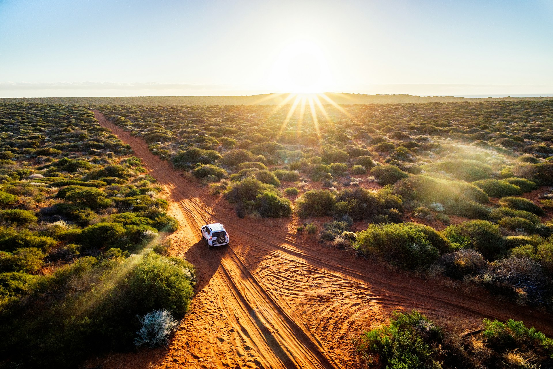 An aerial shot of a white car on a red-dust road heading into the outback