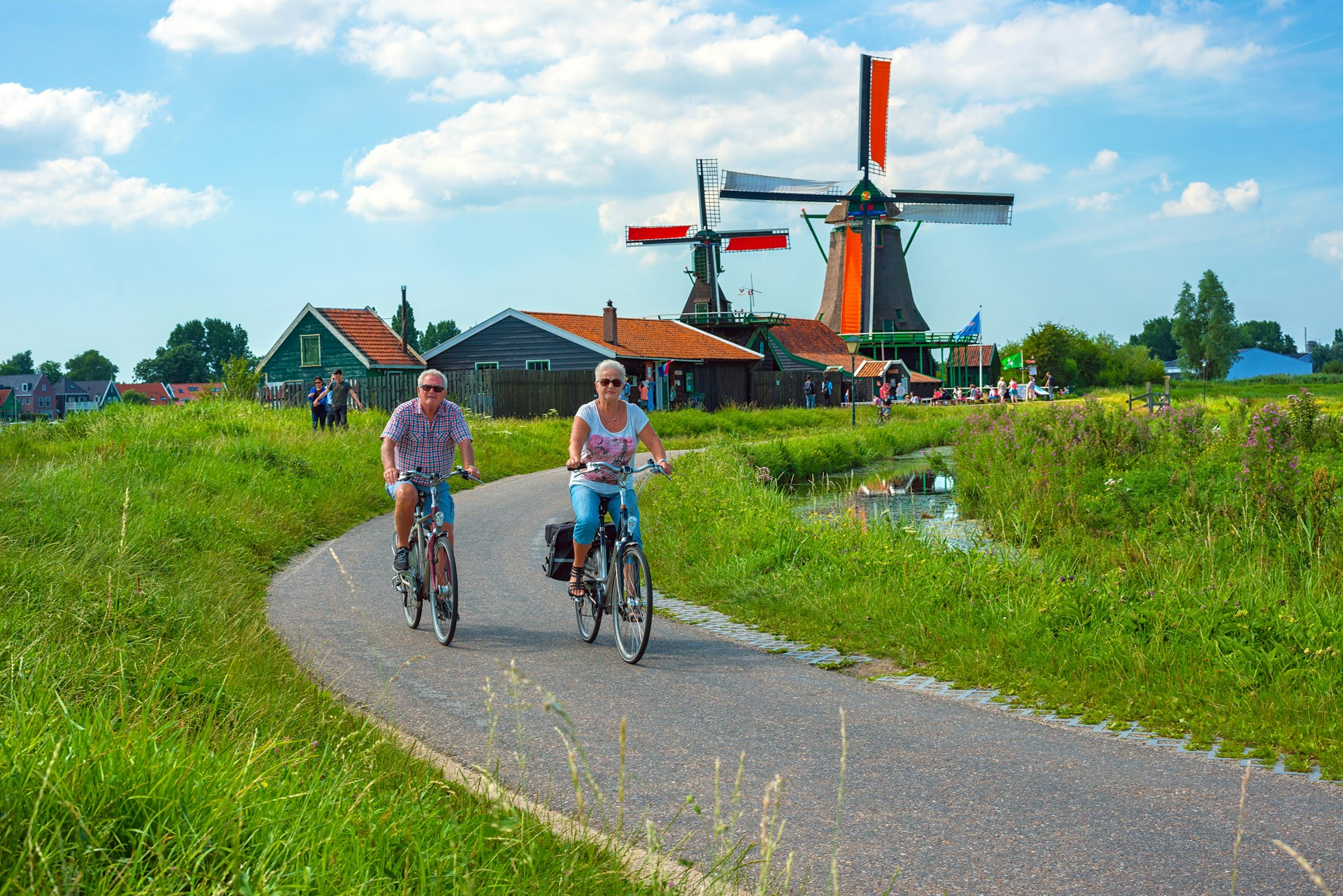 Two cyclists ride bikes on a path surrounded by grassland. There are windmills in the background