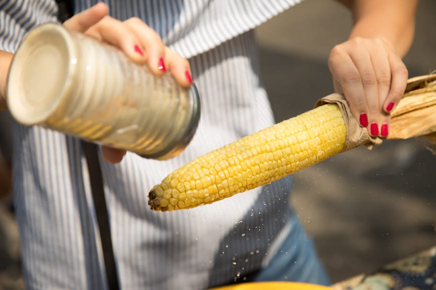 Close-up of a woman holding roasted yellow Mexican corn and shaking seasoning filled with garlic chilli salt