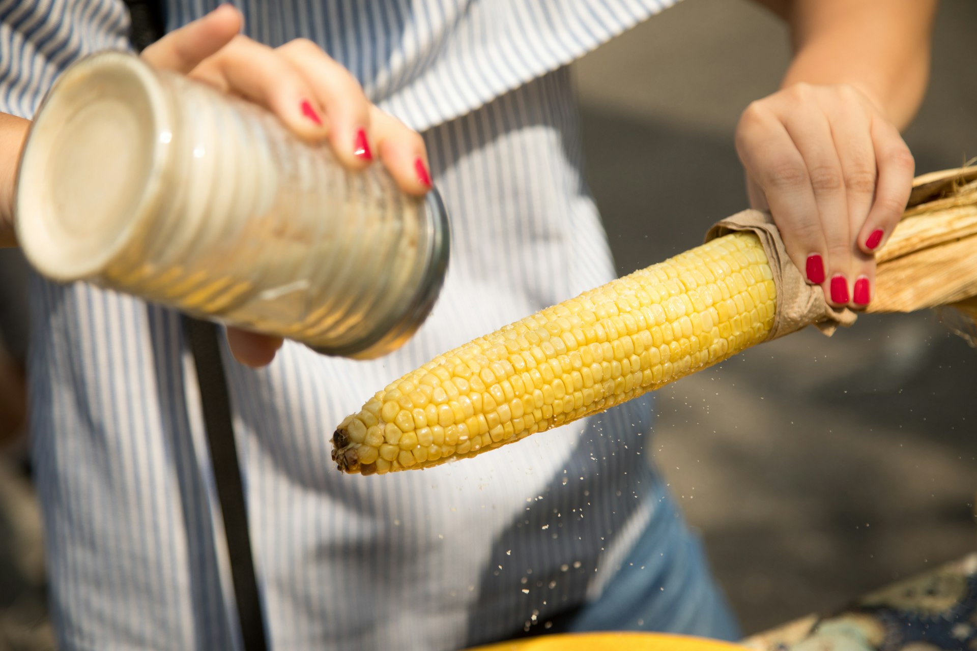 Close-up of a woman holding roasted yellow Mexican corn and shaking seasoning filled with garlic chilli salt