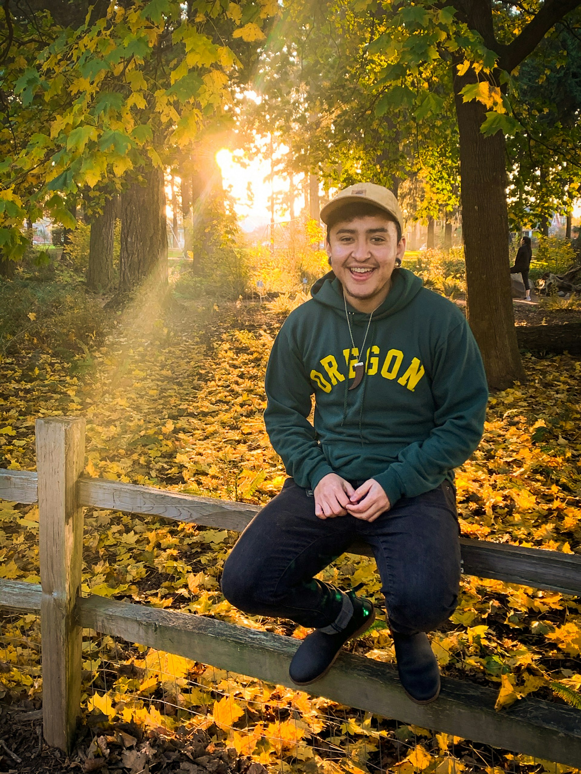 A person sits on a wooden fence smiling at the camera. They are in woodland, with golden sunlight shining through the trees