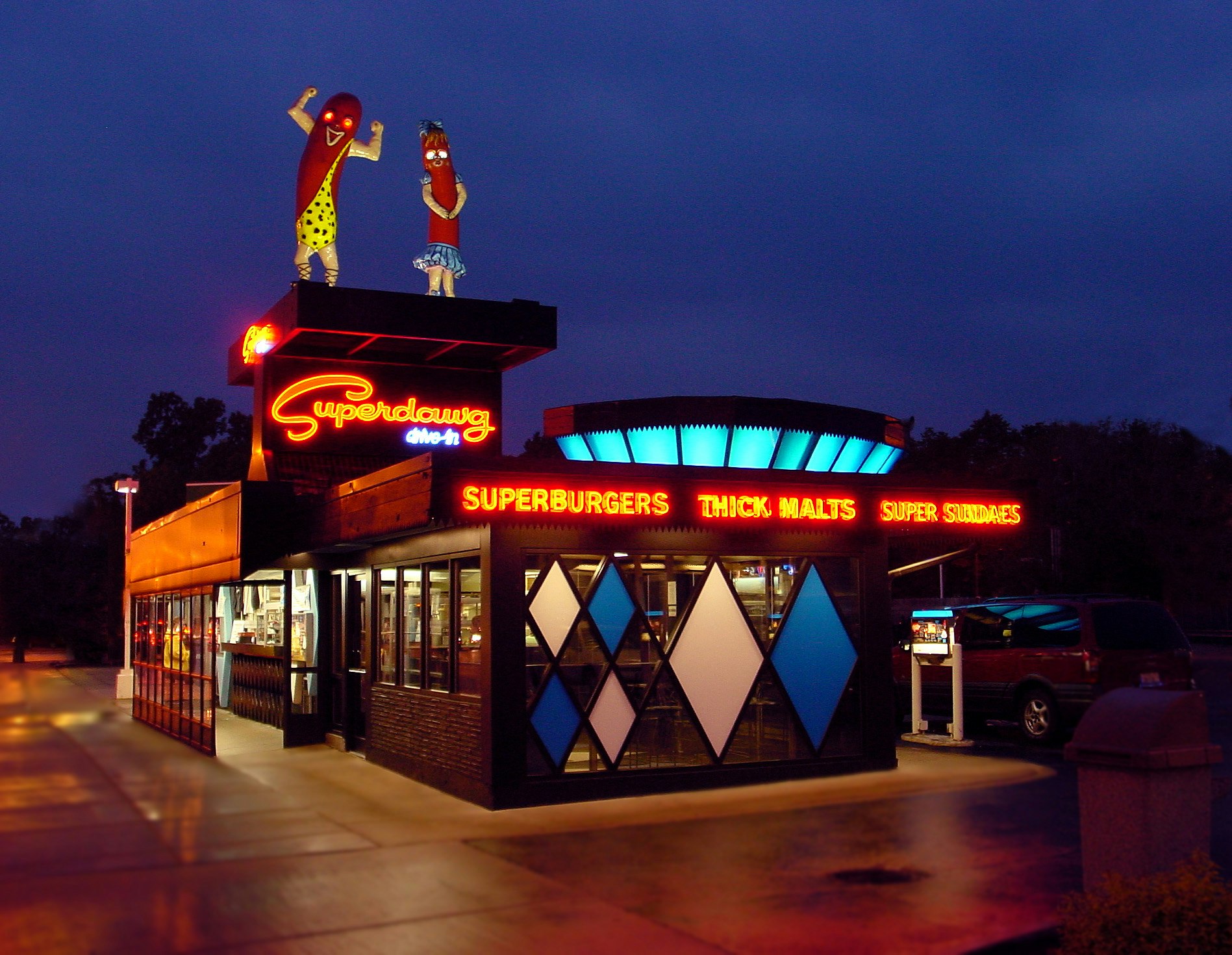 Nattfoto av utsidan av SuperDawg Drive-In.  Högst upp på restaurangtaket står ett par stora glasfiberkorvar