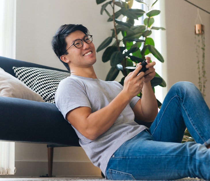 Cheerful young man playing video game, sitting on the floor at home during the day.