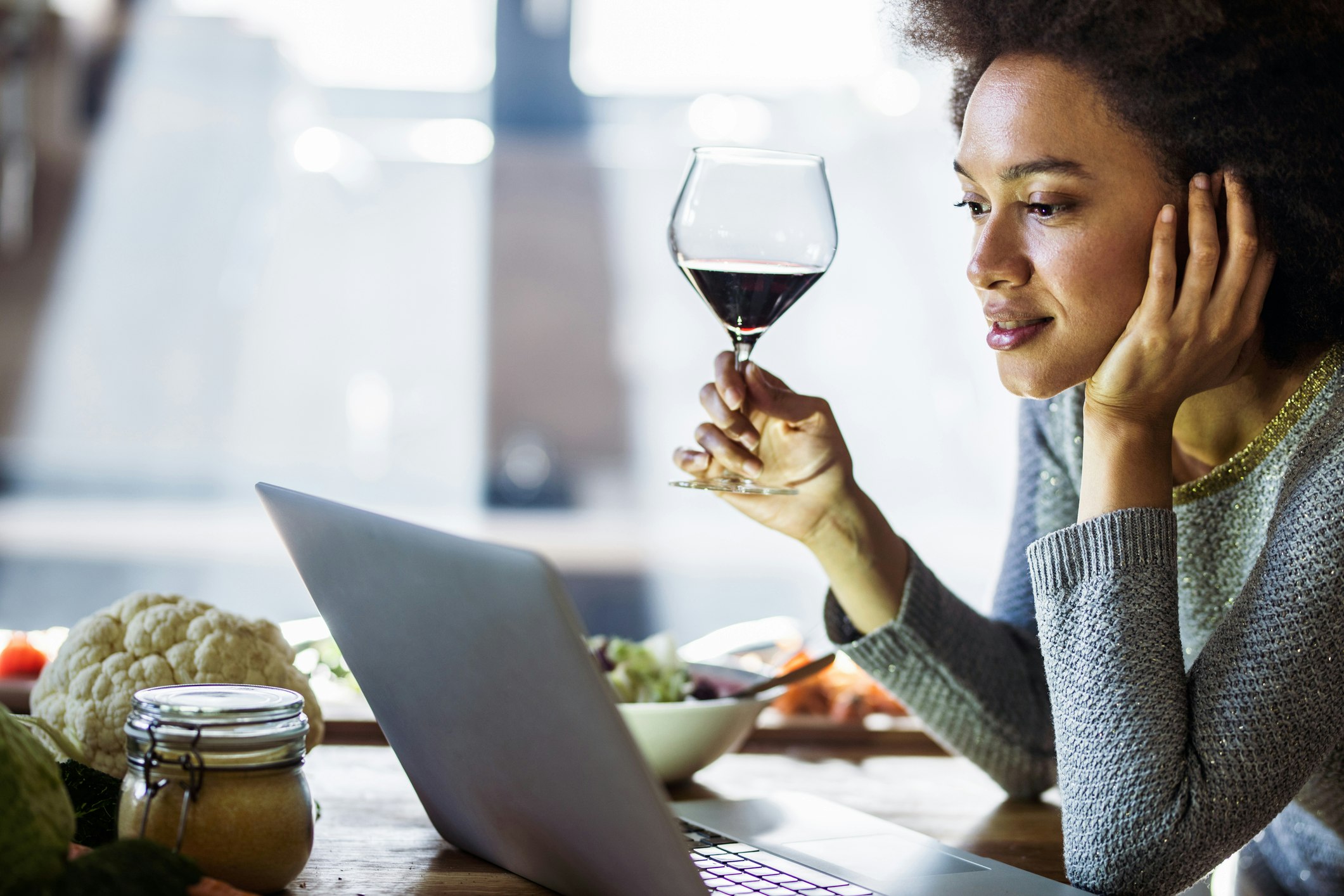 Smiling black woman drinking wine while reading online recipes in the kitchen.
