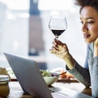 Young African American woman drinking wine while reading recipes over laptop in the kitchen.