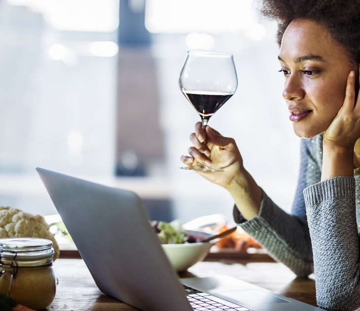 Young African American woman drinking wine while reading recipes over laptop in the kitchen.
