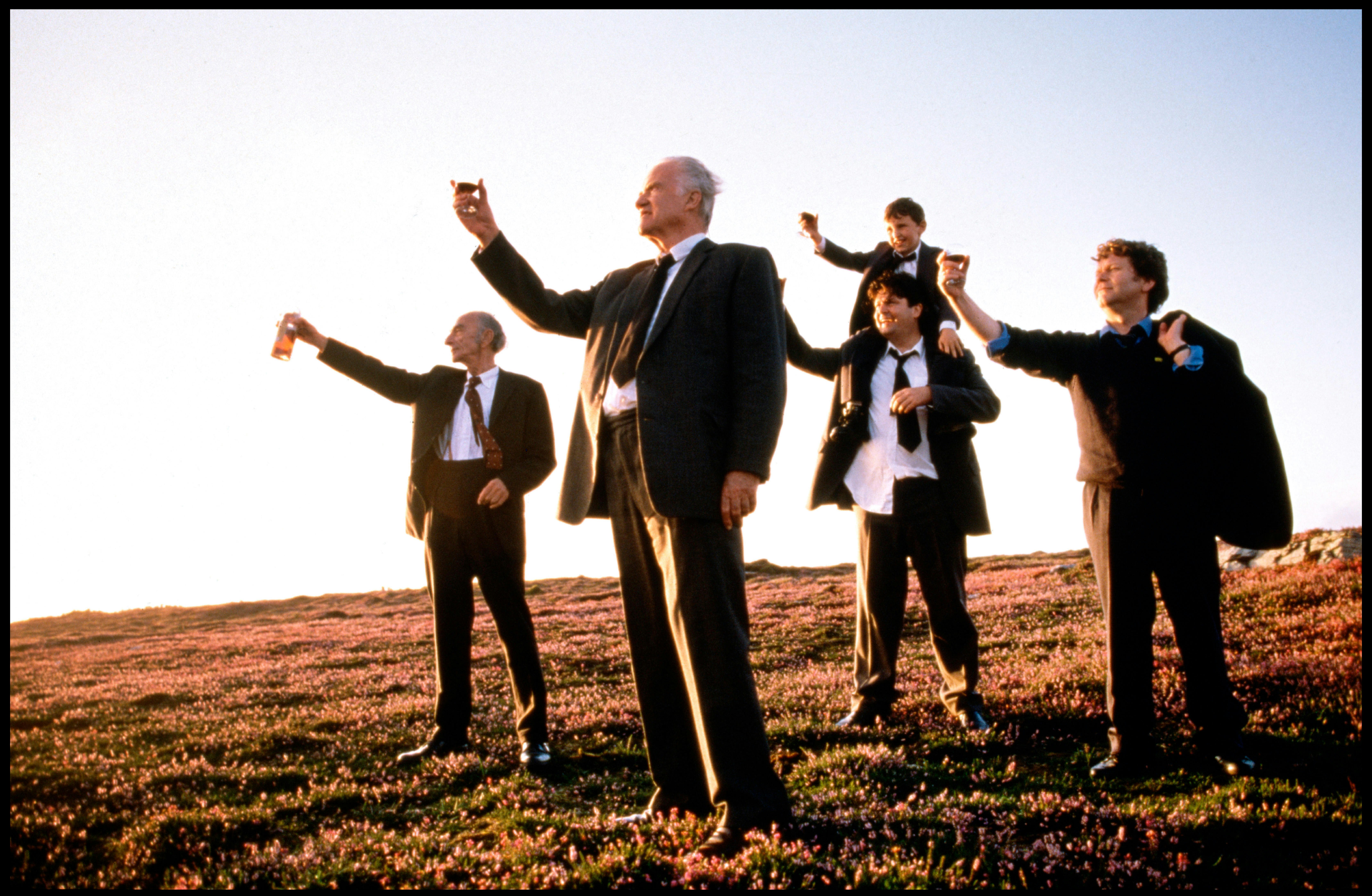 Four men and a boy raise a glass to the sky in the middle of a field. They are all wearong suit and looking in the same direction
