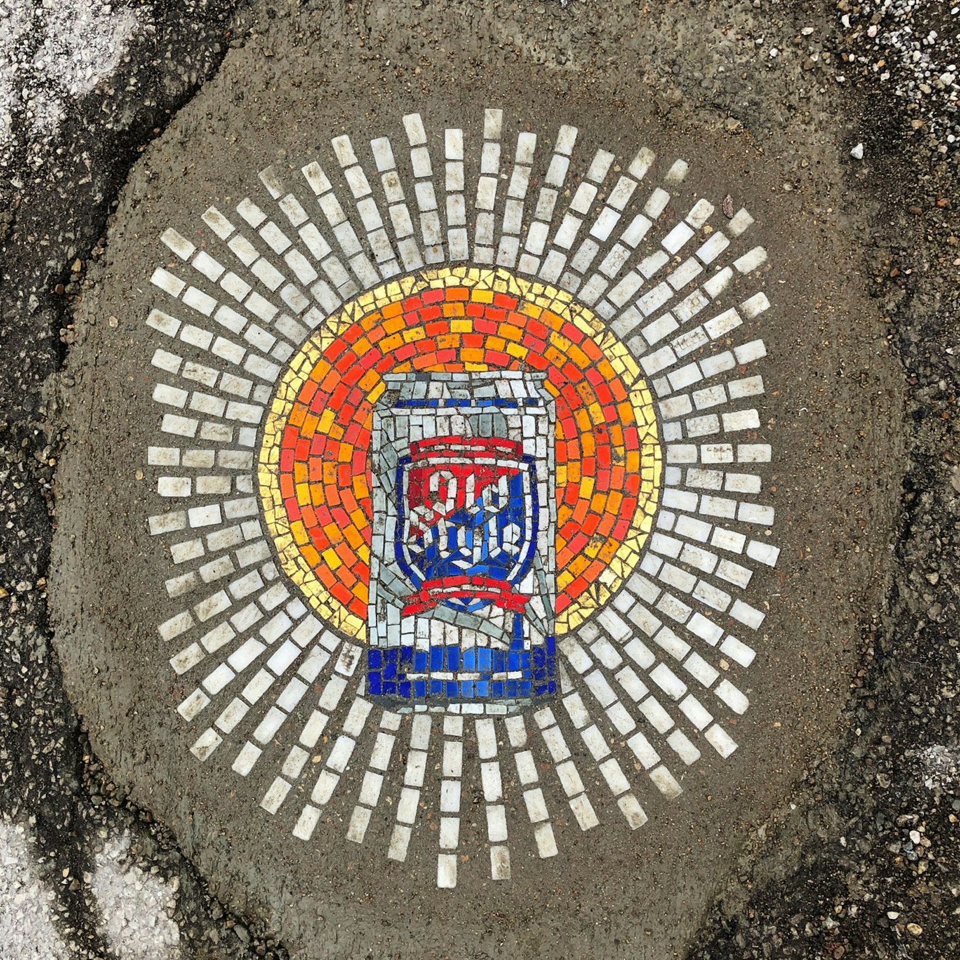 A mosaic depicting a red, blue, and silver can of Old Style lager is surrounded by an orange and yellow hallow and white lines of light in the pavement of a Chicago pothole