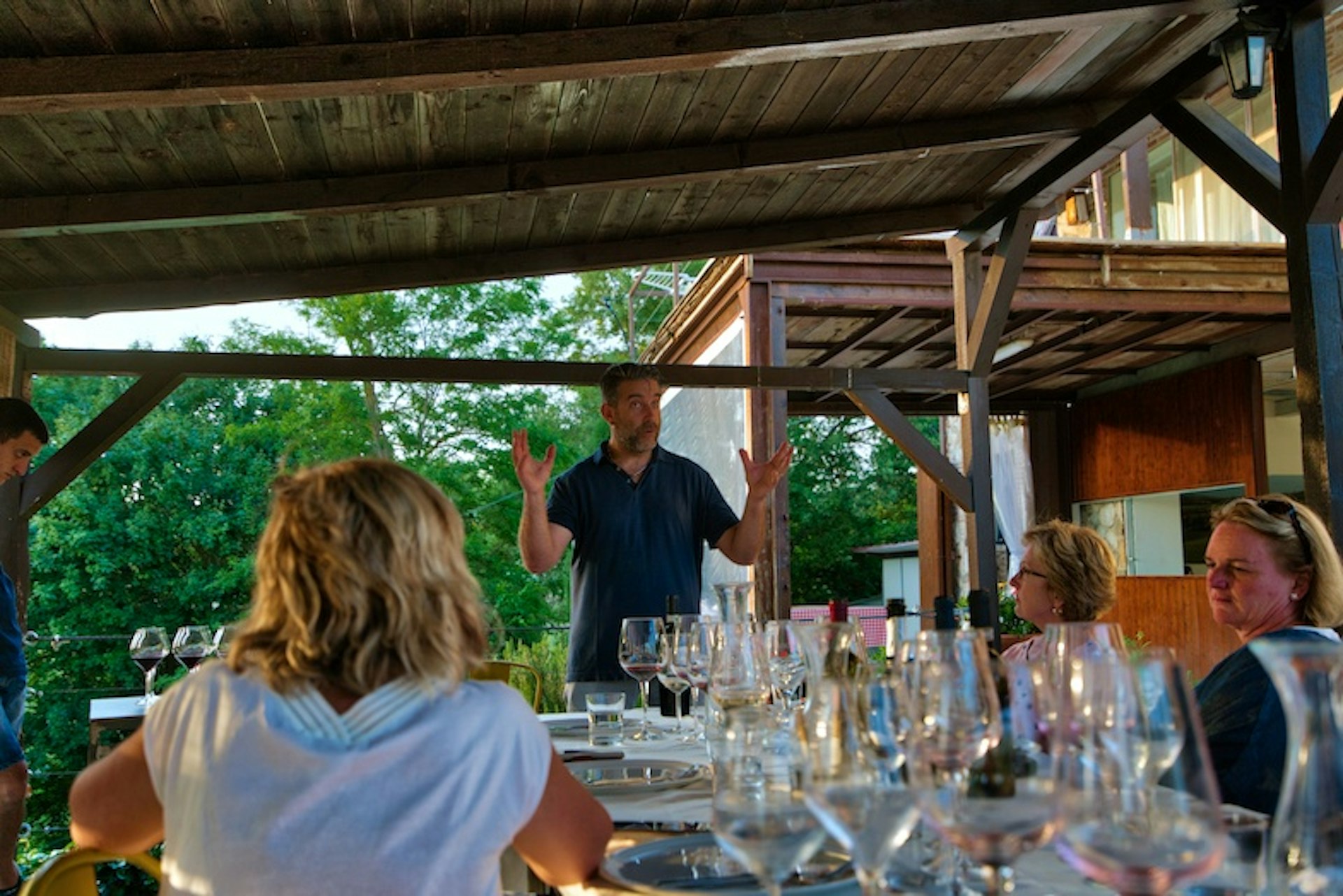 A man stands at the head of a table, arms open, while people sit around. The table is covered in wine glasses and a white tablecloth.