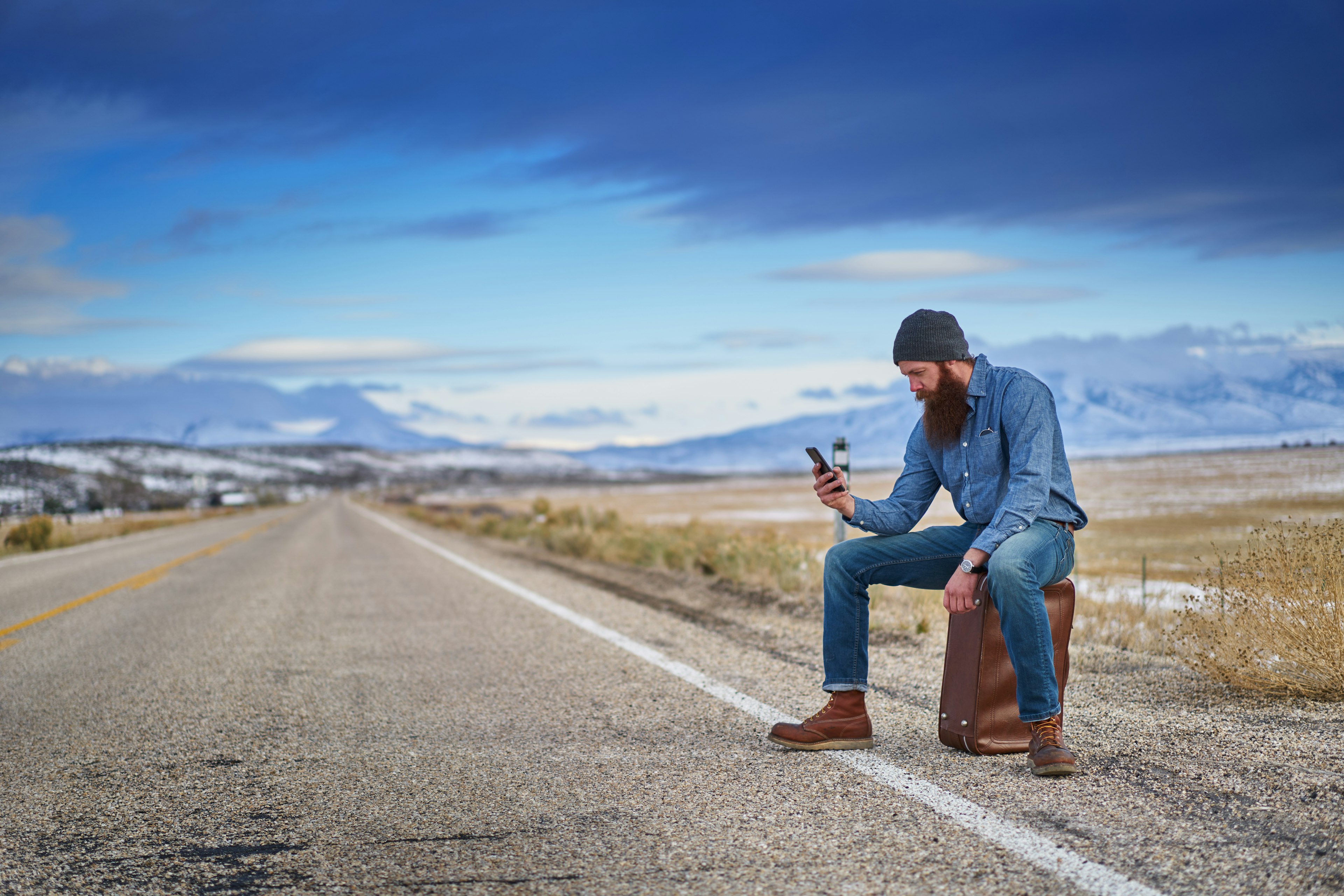 A bearded hitch hiker sitting on a suitcase and using a smart phone on the side of deserted road.