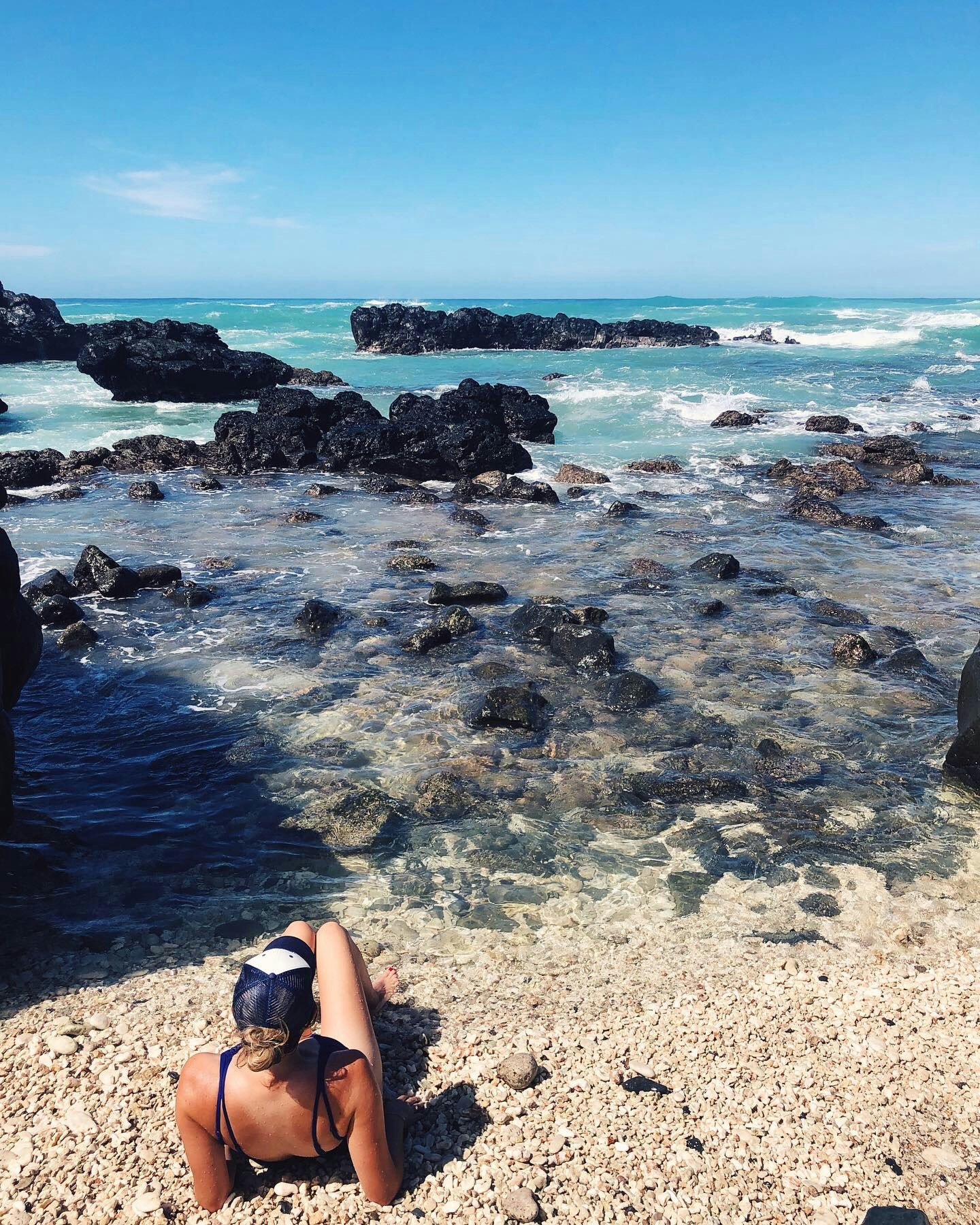 A woman in a hat and swimwear faces away from the camera. She's sat on the beach where the sea meets the sand