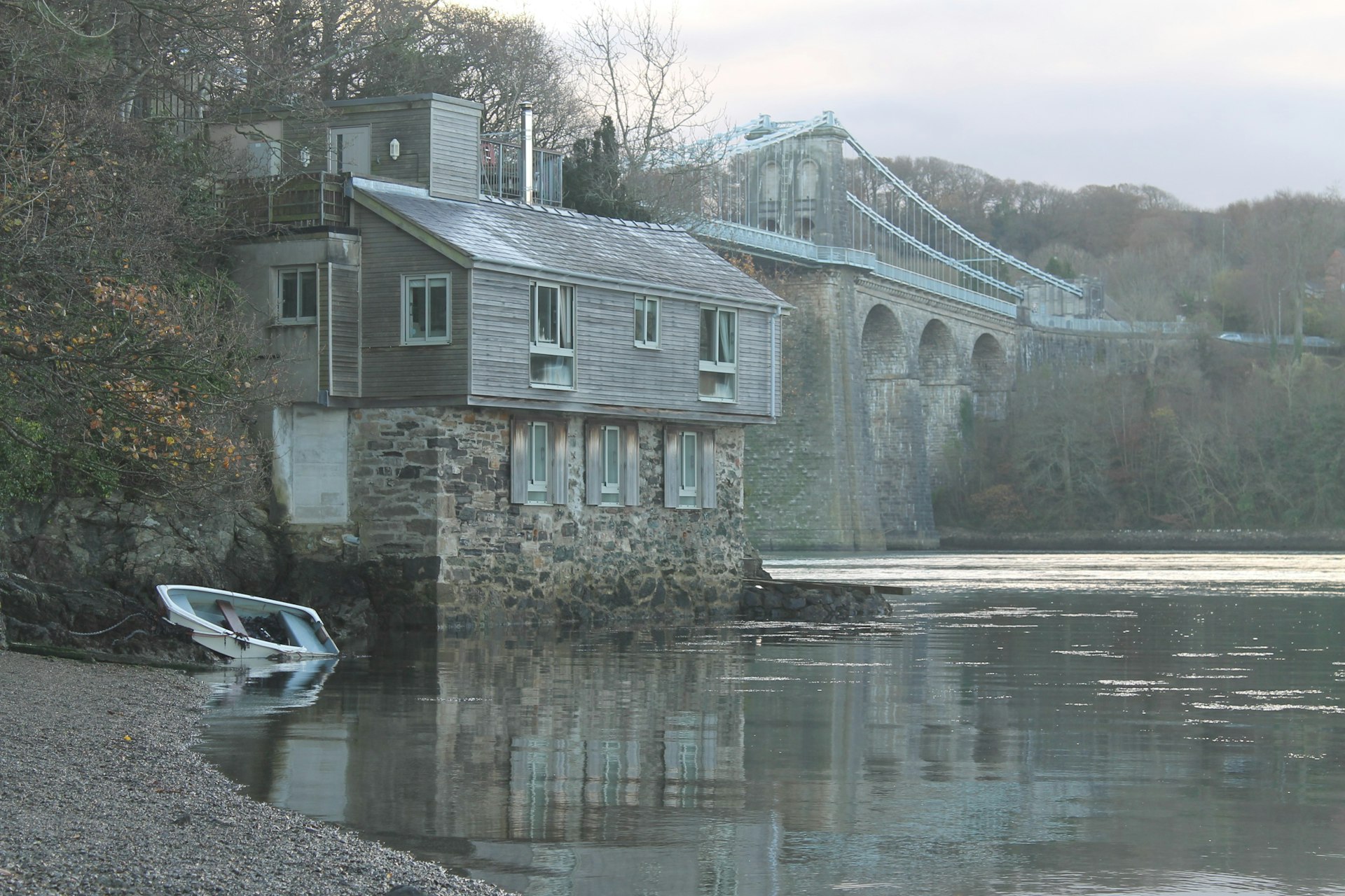 A boat house stands on the edge of the water at the end of a walkway in Anglesey. Behind it, a large bridge is visible.