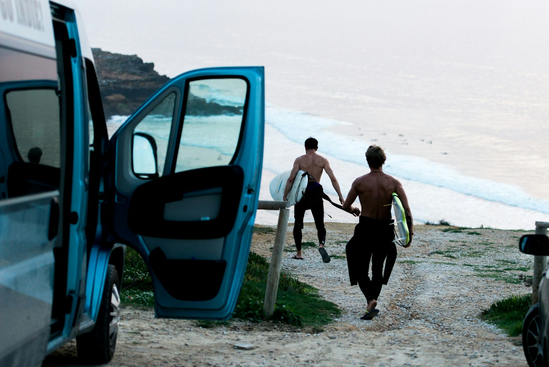 Two men with surfboard heading into the sea next to a campervan 