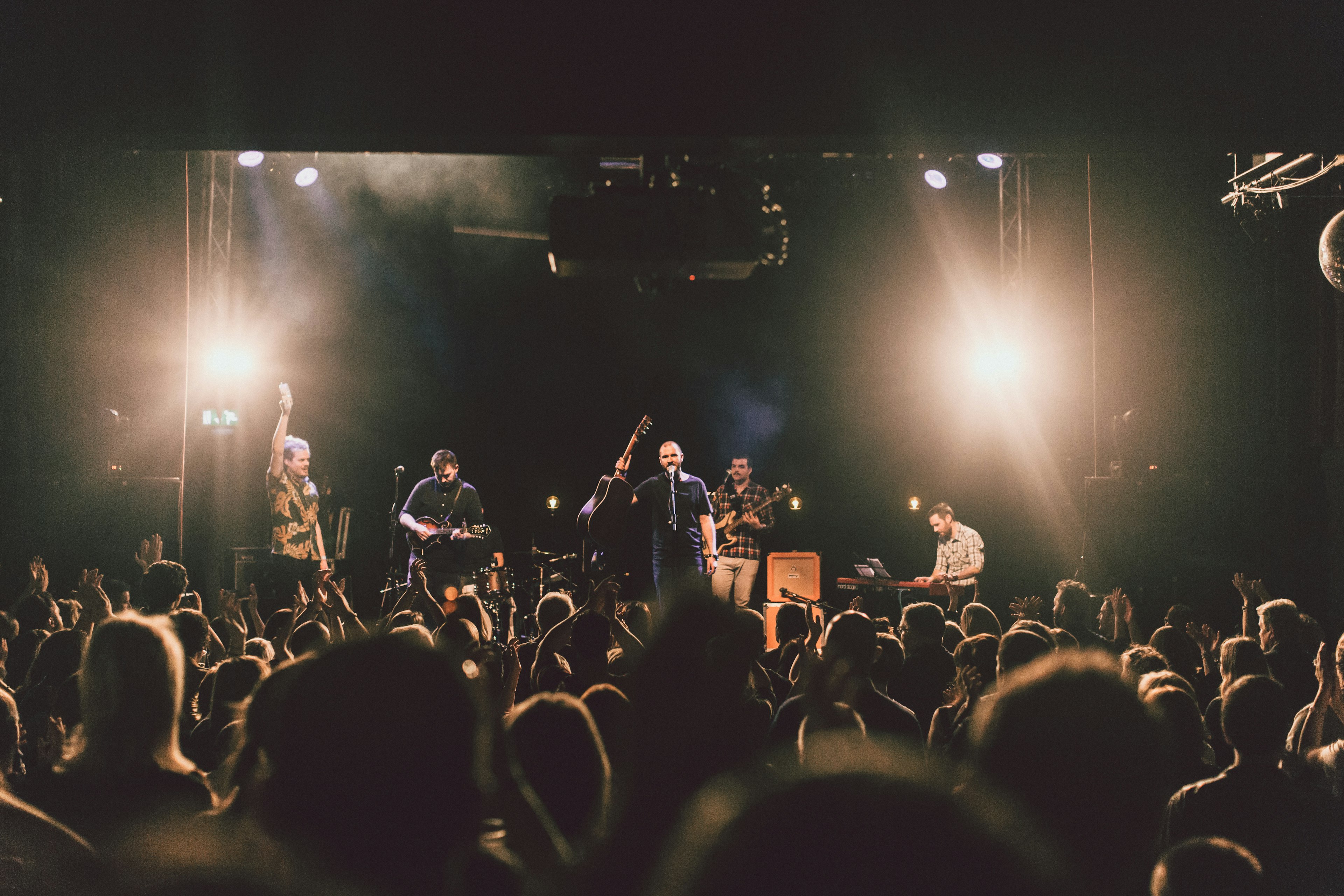 A band performing on stage. The man in the center is holding a guitar to his side and speaking into the microwave to the large crowd