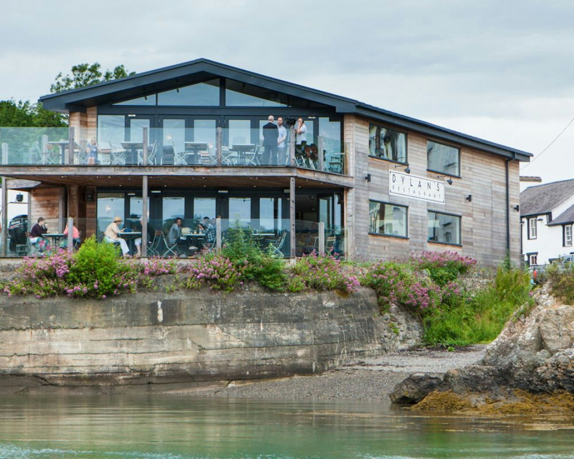 A view of Dylan's restaurant in Menai Bridge, Anglesey, from the water. The restaurant has a balcony, on which a number of diners are sitting, that overlooks the calm water.