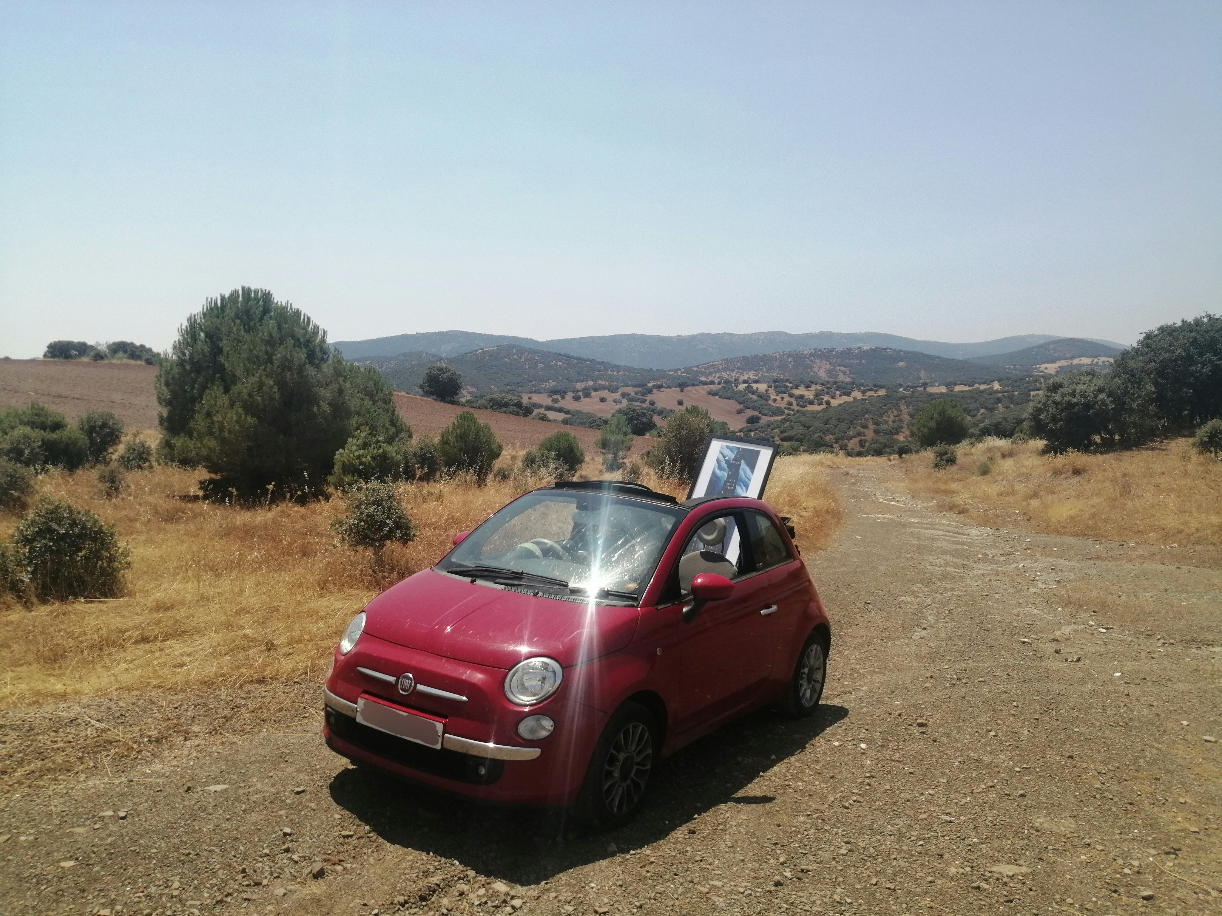 A red Fiat 500 convertible car on a dusty road in the sunshine, with a painting sticking out through the roof 