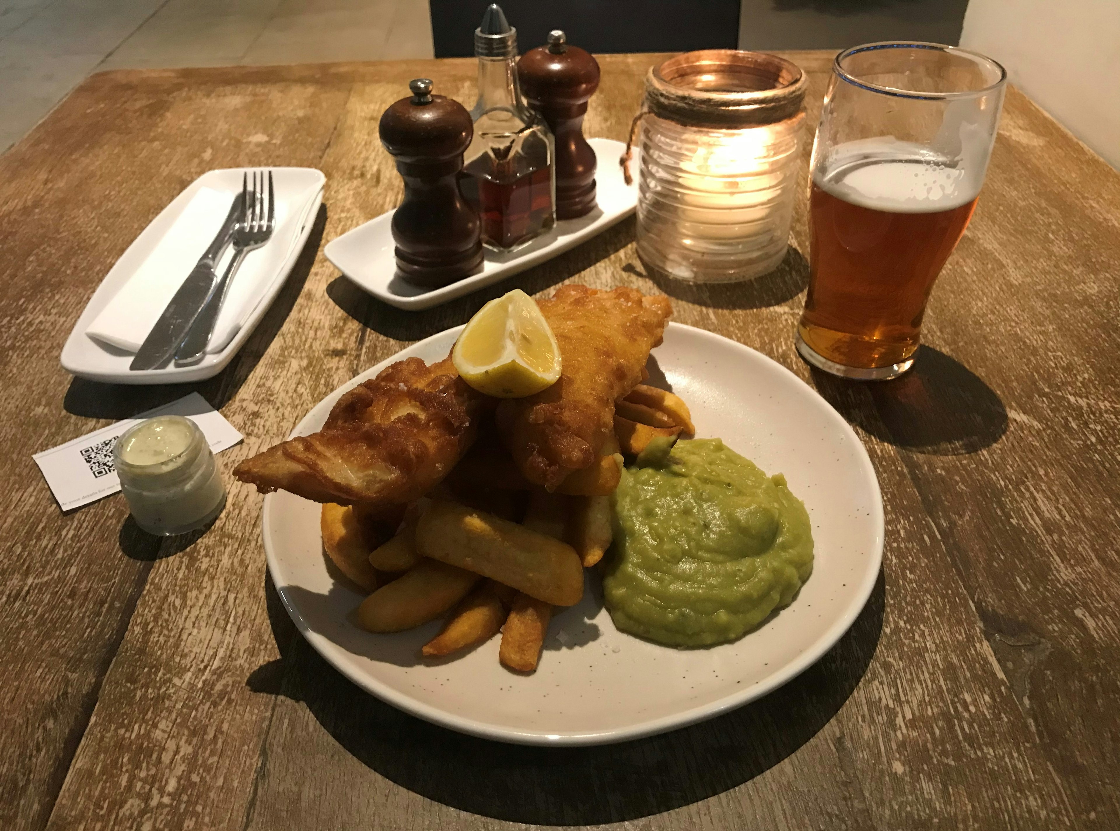 A plate of fish and chips, with a large dollop of mushy peas, stands on a wooden table, with condiments and a pint of beer around it.