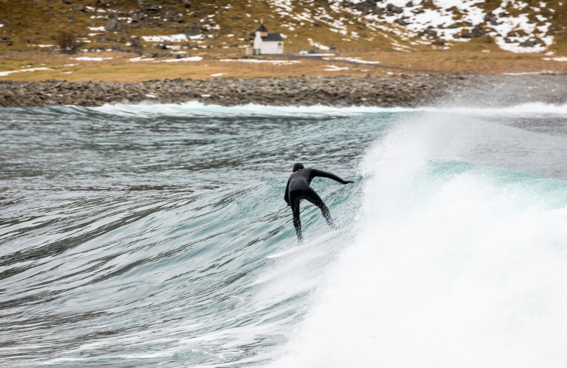 A surfer in full gear covering them head to toe surfs a wave at a bay. There is snow on the rocks of the beach.