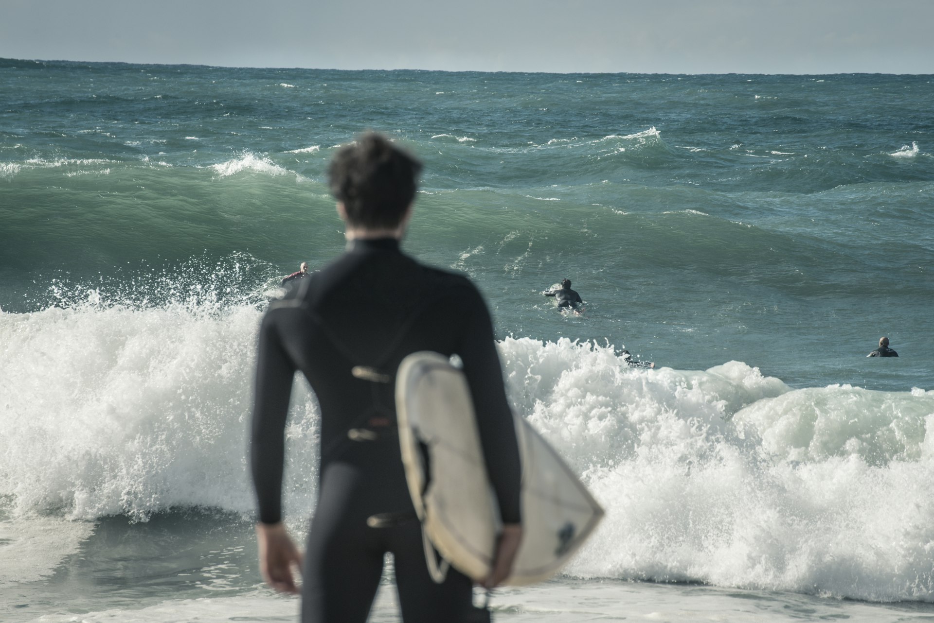 As costas de um surfista segurando uma prancha e olhando para o mar, para outros surfistas se aproximando de ondas grandes