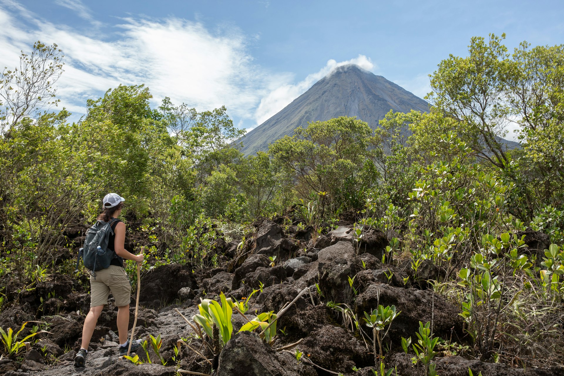 A hiker with a stick pauses on a trail to gaze at the huge volcano that dominates the jungle landscape