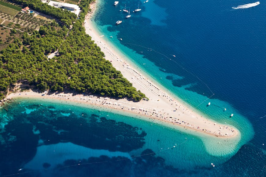 Aerial photograph of Zlatni Rat beach on Brac Island. The beach is distinctive as it forms a sharp triangle of white sand at the end of the island. The beach is surrounded by blue water with many people sunning themselves on the sand.