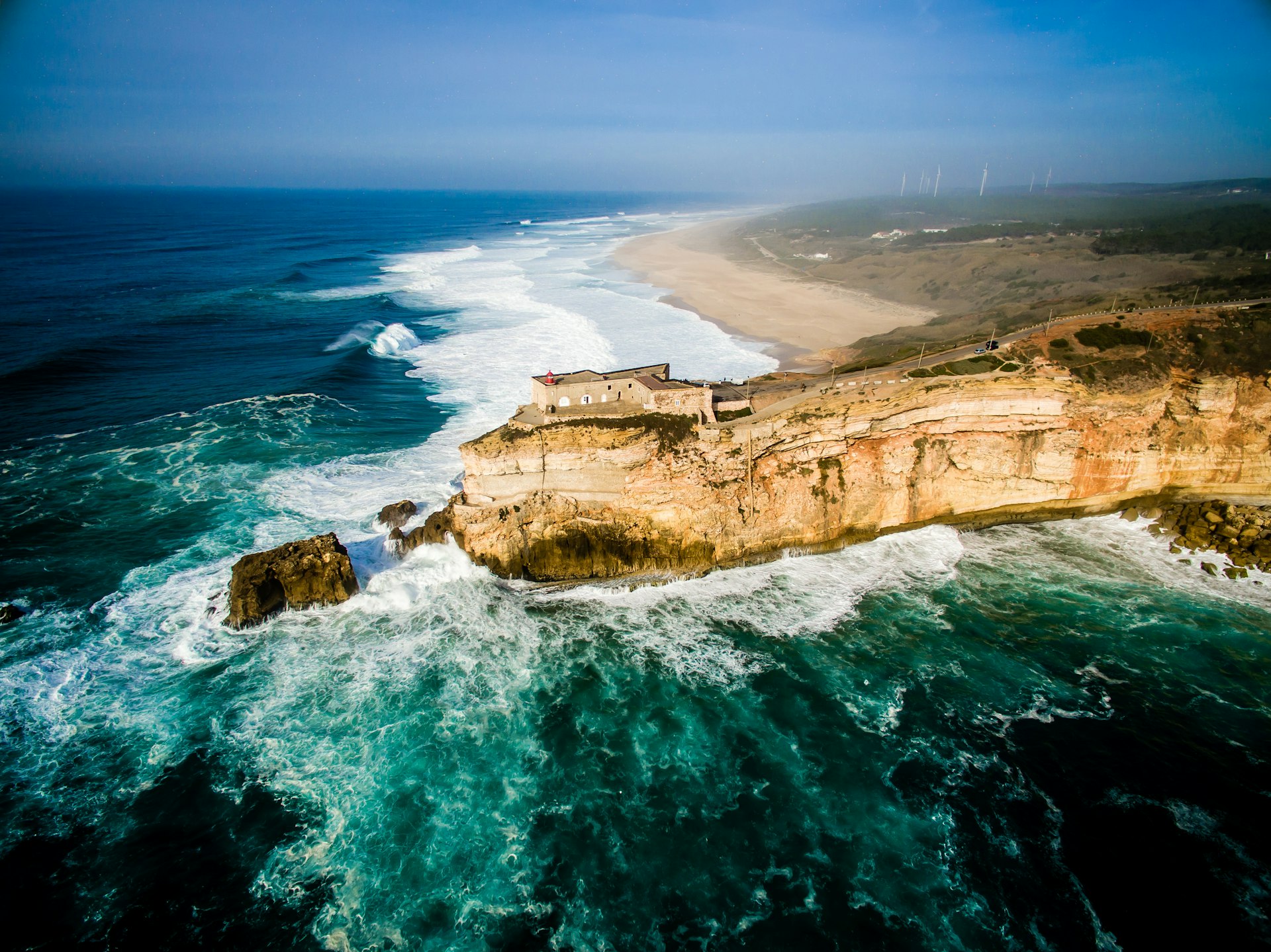 Aerial shot of a narrow peninsula with a clifftop building surrounded by rough blue ocean