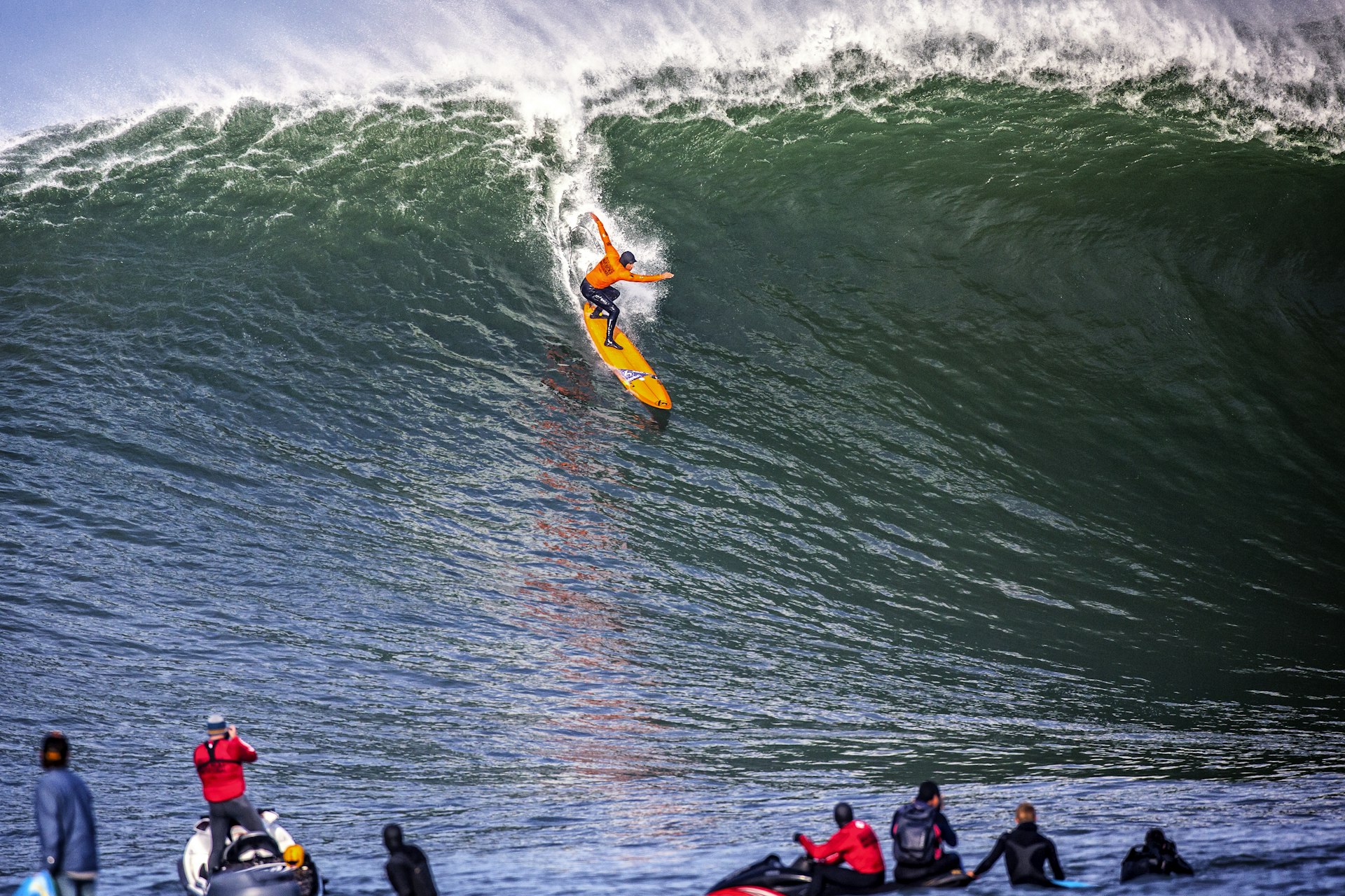 A surfer tops a wave heading towards a series of onlookers sat on jet skis