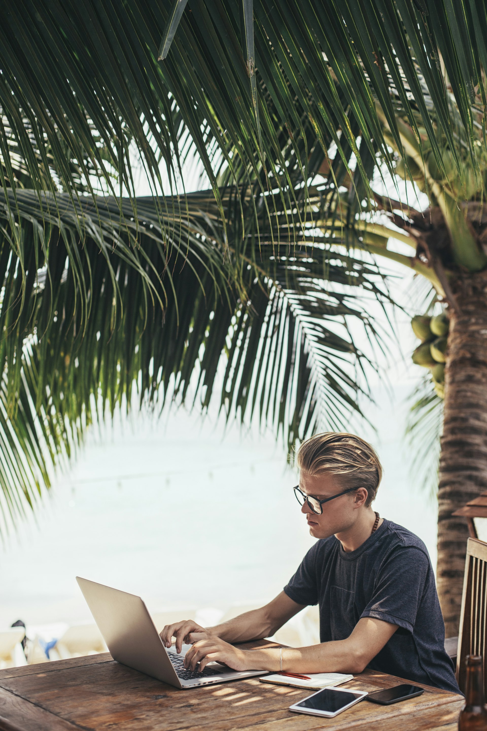 A white man using a laptop at a table near a palm-fringed beach