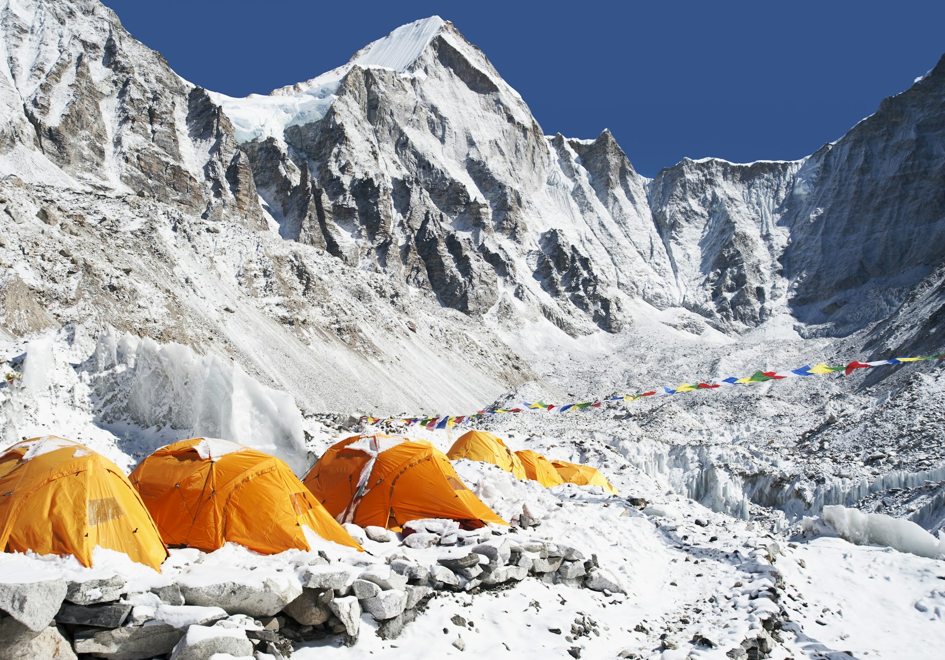 A row of bright-orange tents sit against the white and grey hulks of Everest Base Camp as colourful prayer flags flap in the wind behind 