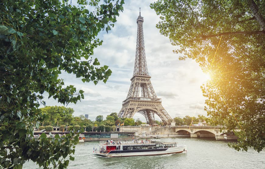 A boat cruises along a wide river towards a bridge with the huge Eiffel Tower looming over it